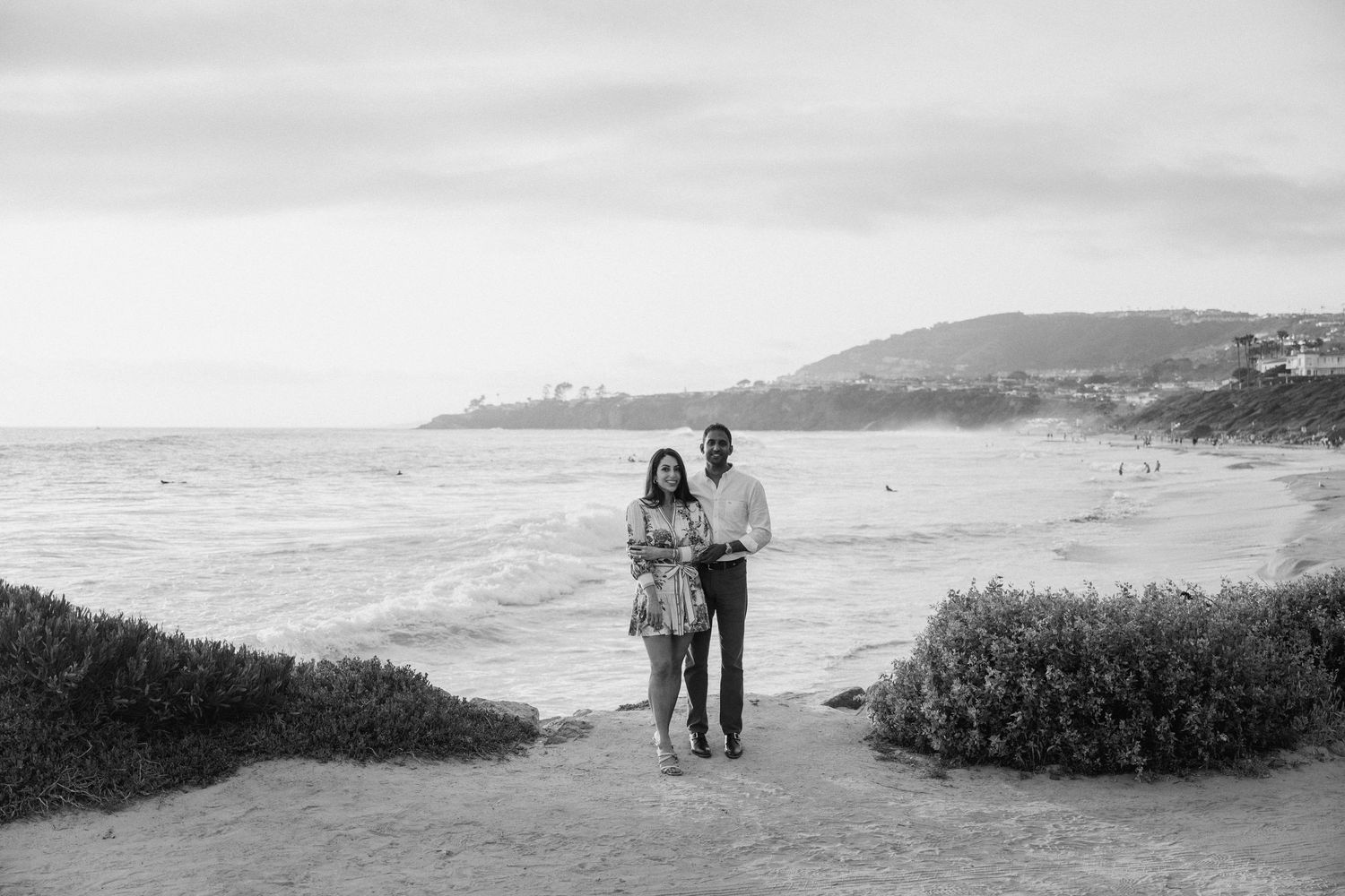 A stunning coastal landscape features a green lawn overlooking a beach with mountains in the distance along the ocean shore.