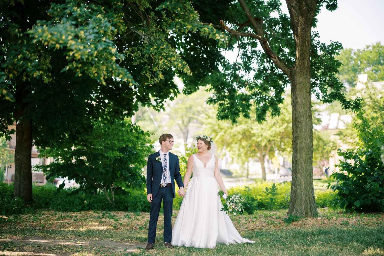 Two people in formal wedding attire stand together under large shady trees in a park setting.