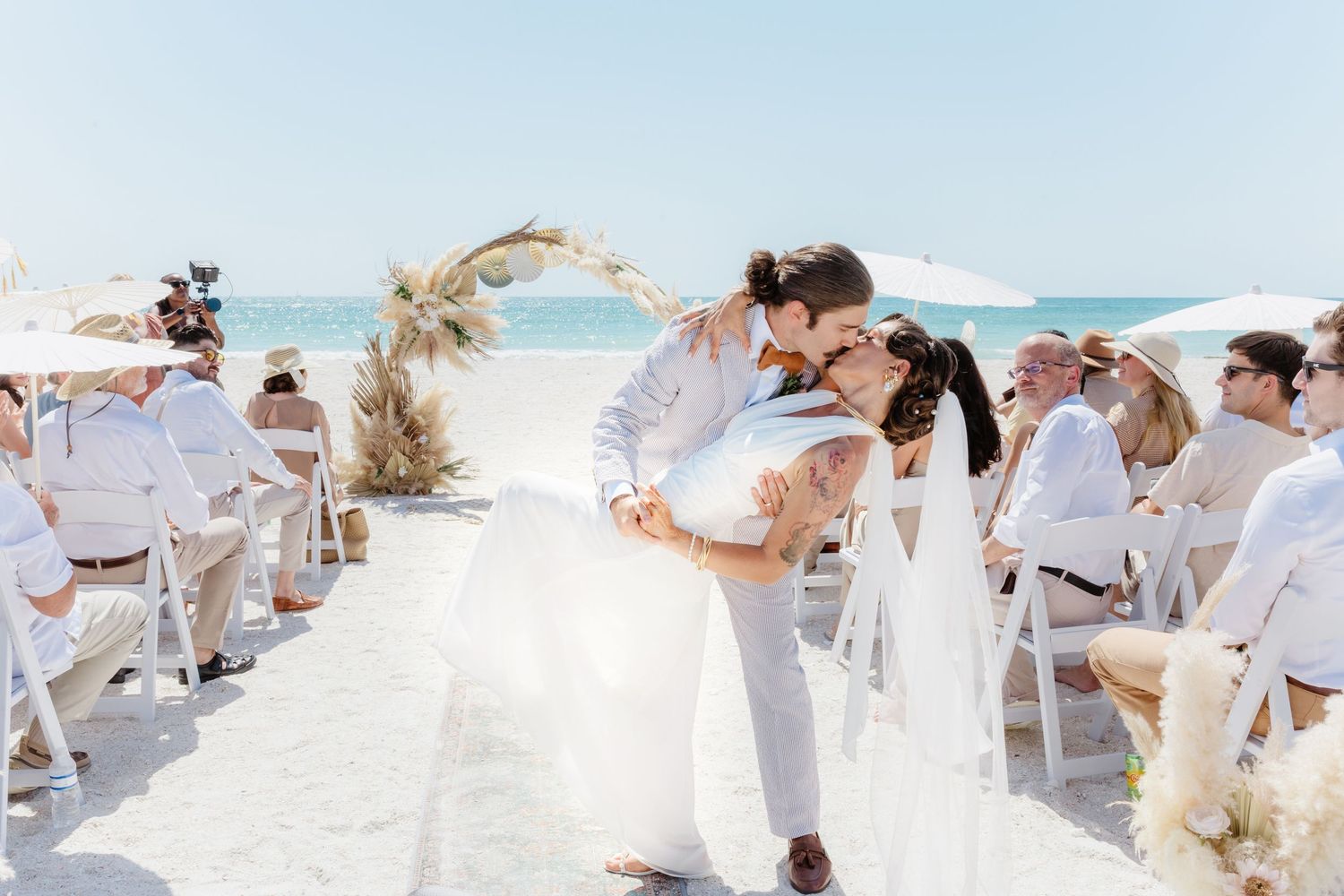 Guests seated at a beachside wedding ceremony with white chairs and a floral arch overlooking the ocean.