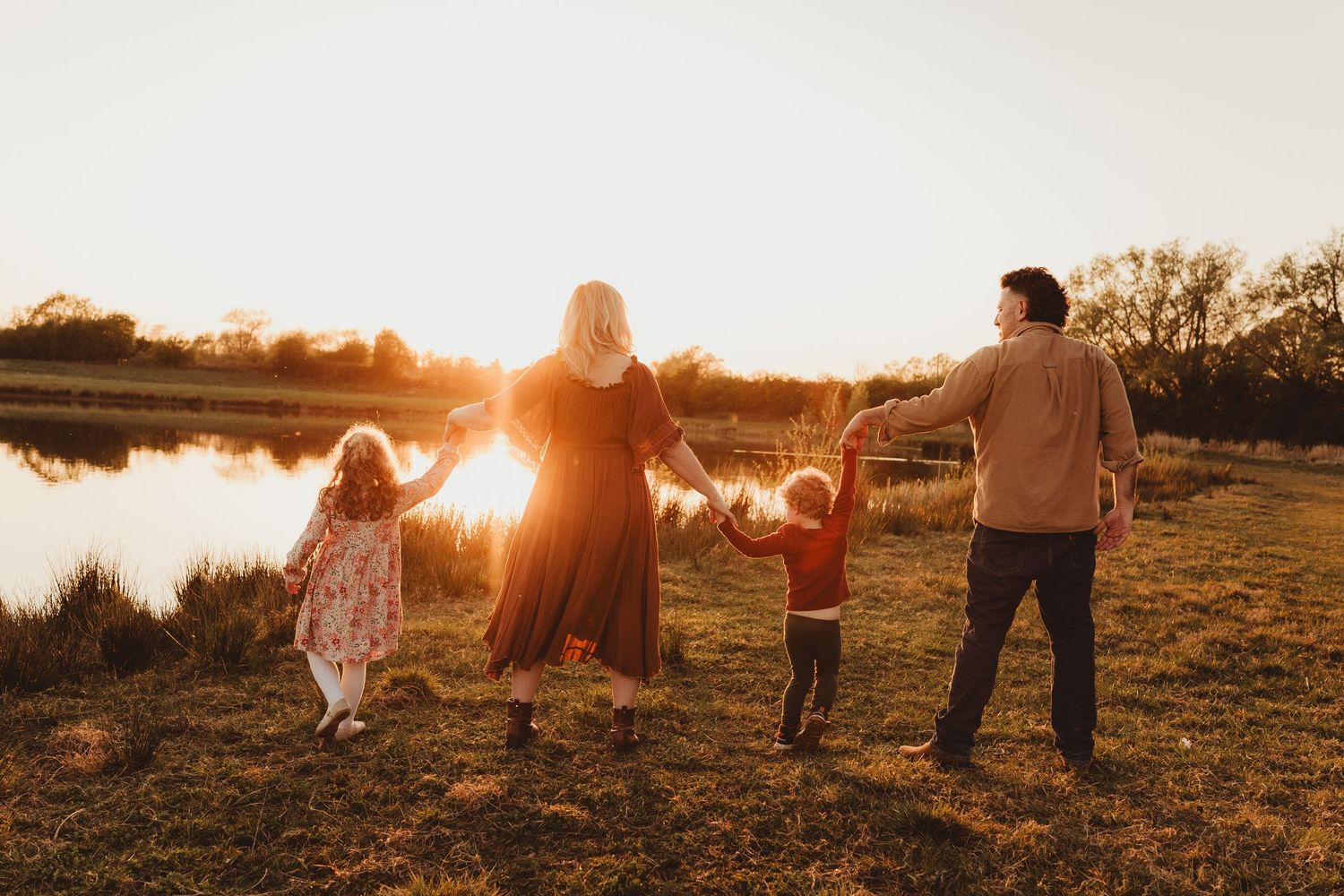 Family enjoys playful moments together by a lake during golden hour sunset with silhouetted trees in background.
