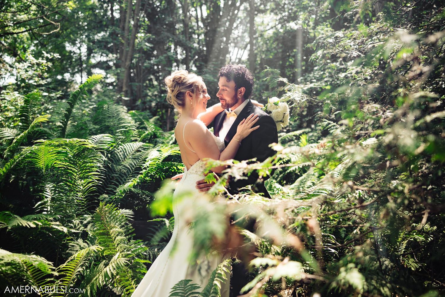 A romantic wedding couple embraces in a lush forest setting surrounded by green ferns and dappled sunlight.