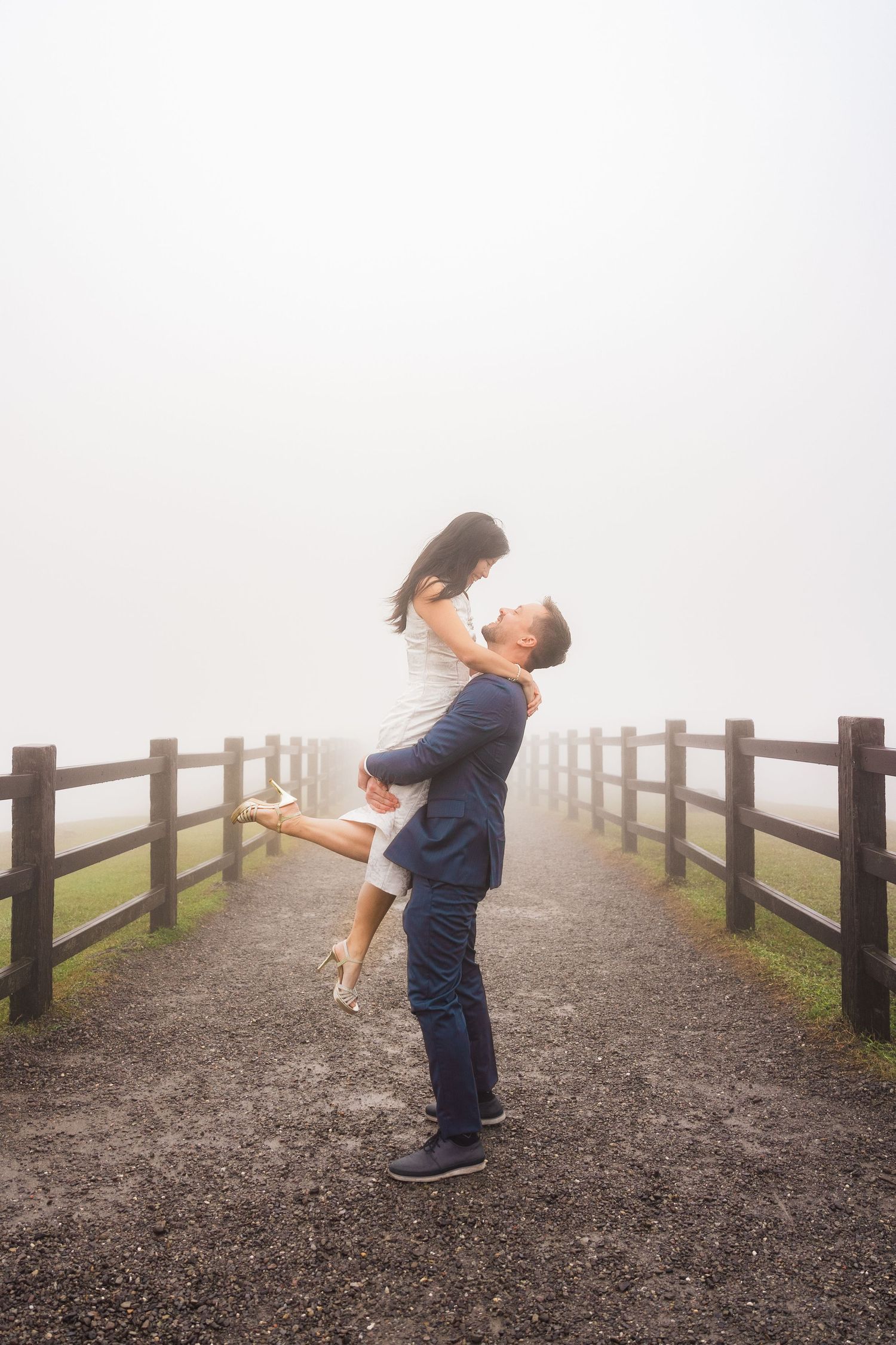 Couple embracing on a misty wooden boardwalk path with a romantic lift pose.