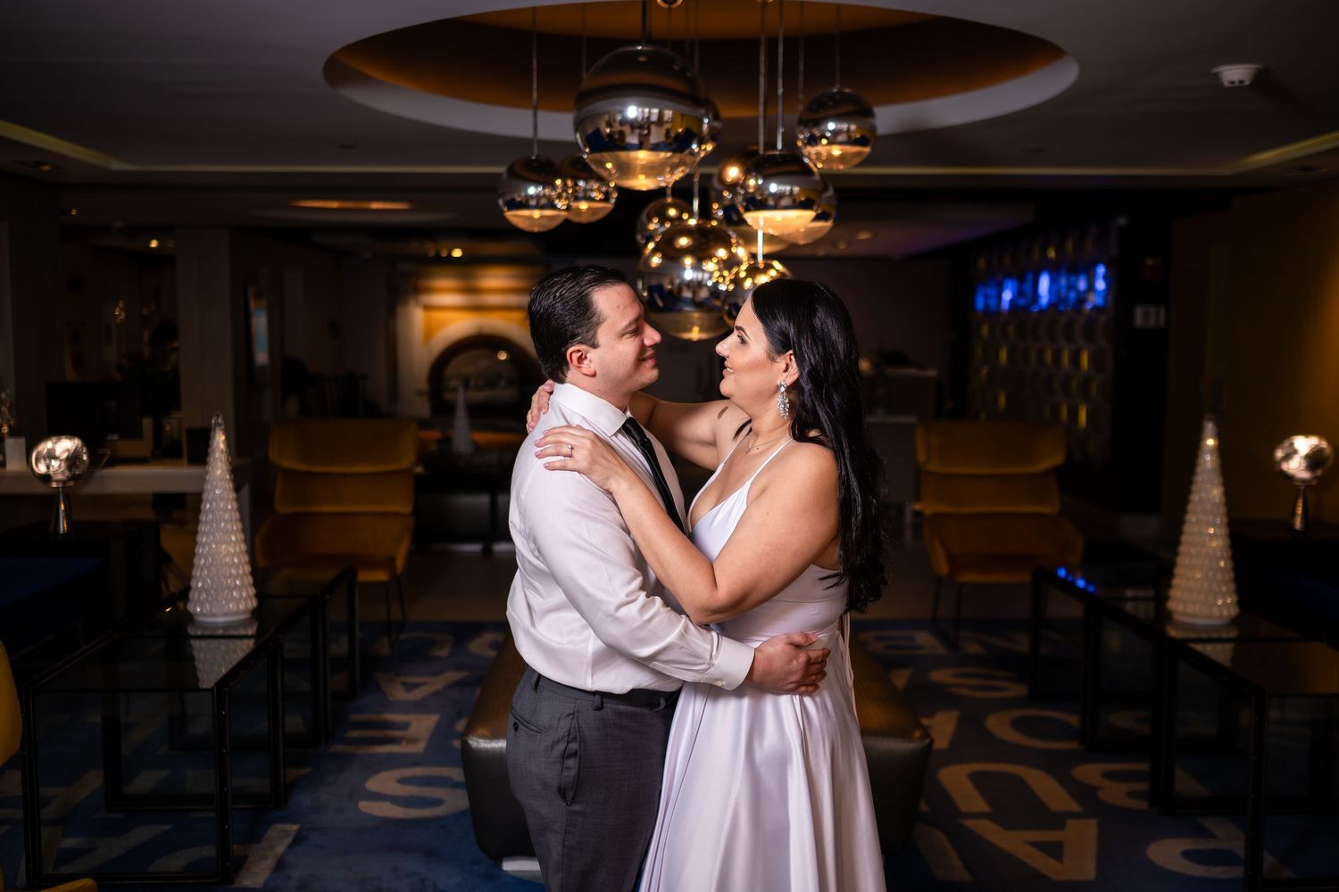 Couple sharing intimate dance moment under elegant chandelier in dimly lit ballroom.