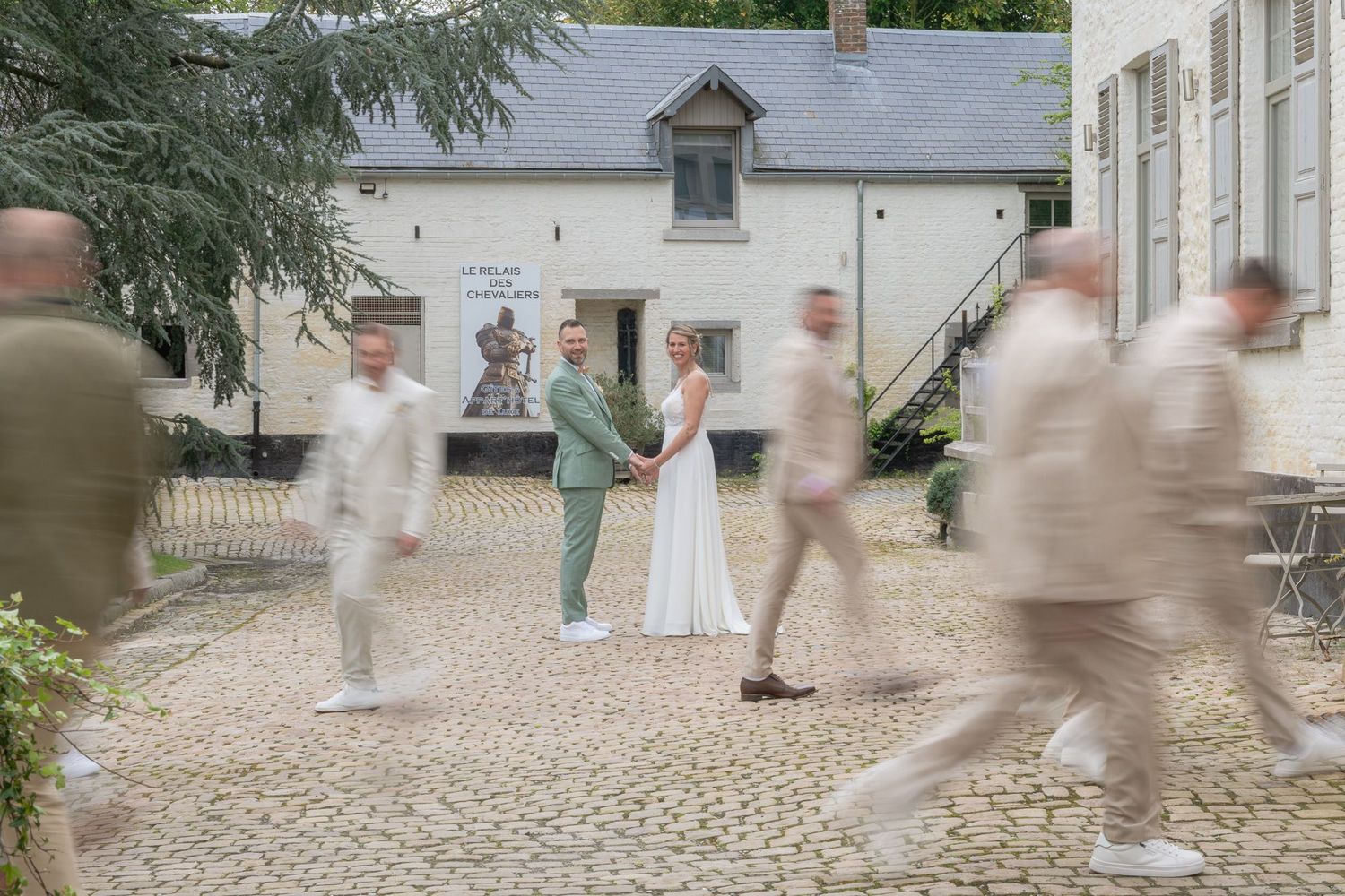 Motion blur captures wedding guests in white attire walking across a cobblestone courtyard outside a traditional building.
