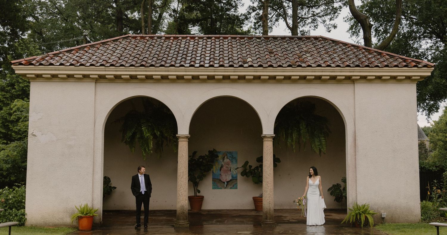 A couple walks along a wet stone path in a lush garden with a small Italian gazebo in the background on their wedding day.