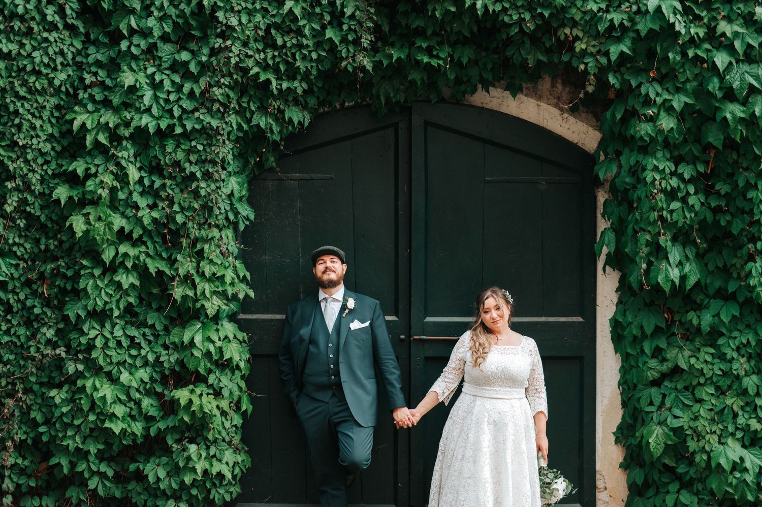 A bride and groom hold hands in front of a large ivy-covered dark green door on their wedding day.