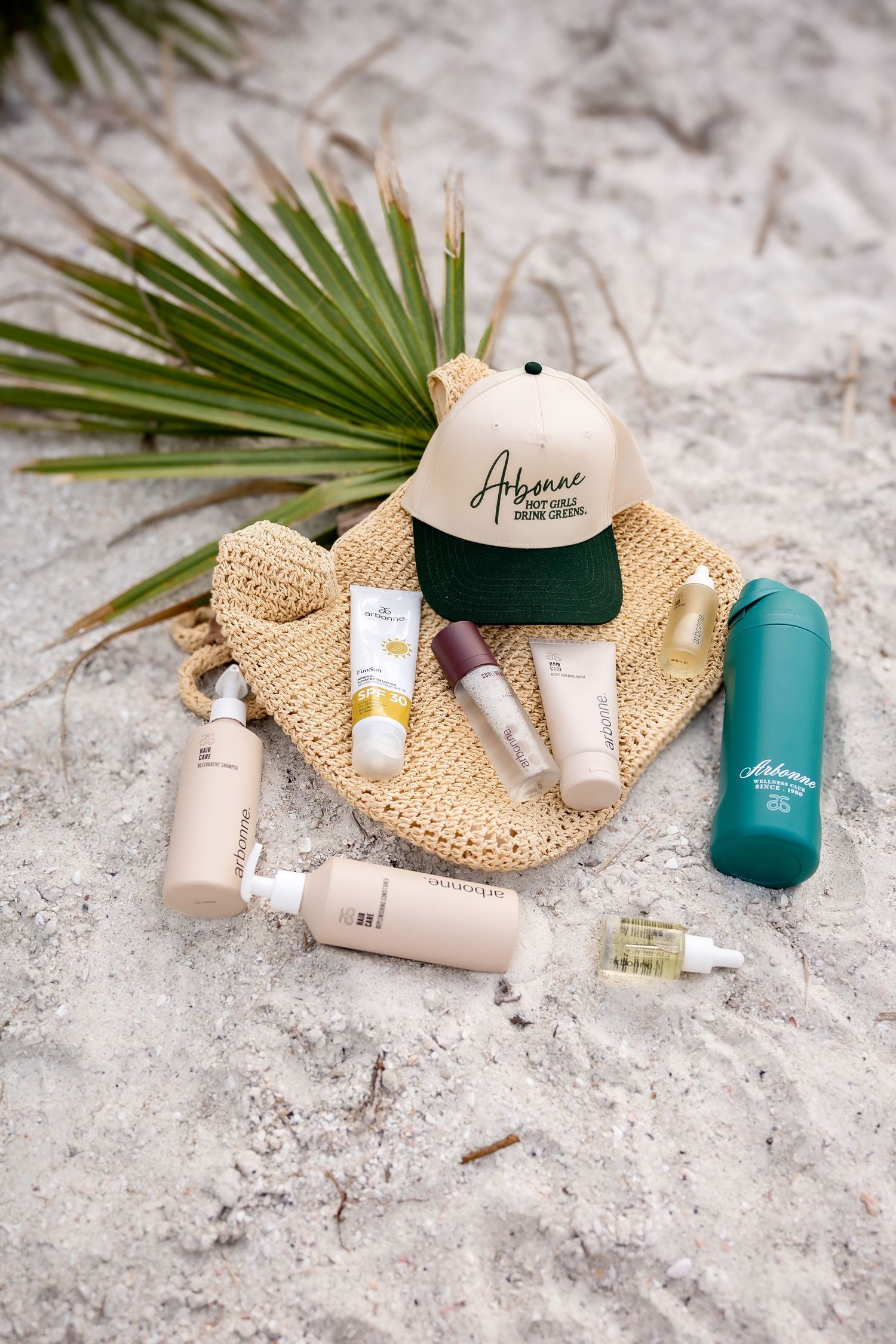 Arbonne skincare products displayed on sand with woven bag, palm leaf, cap, and teal water bottle.