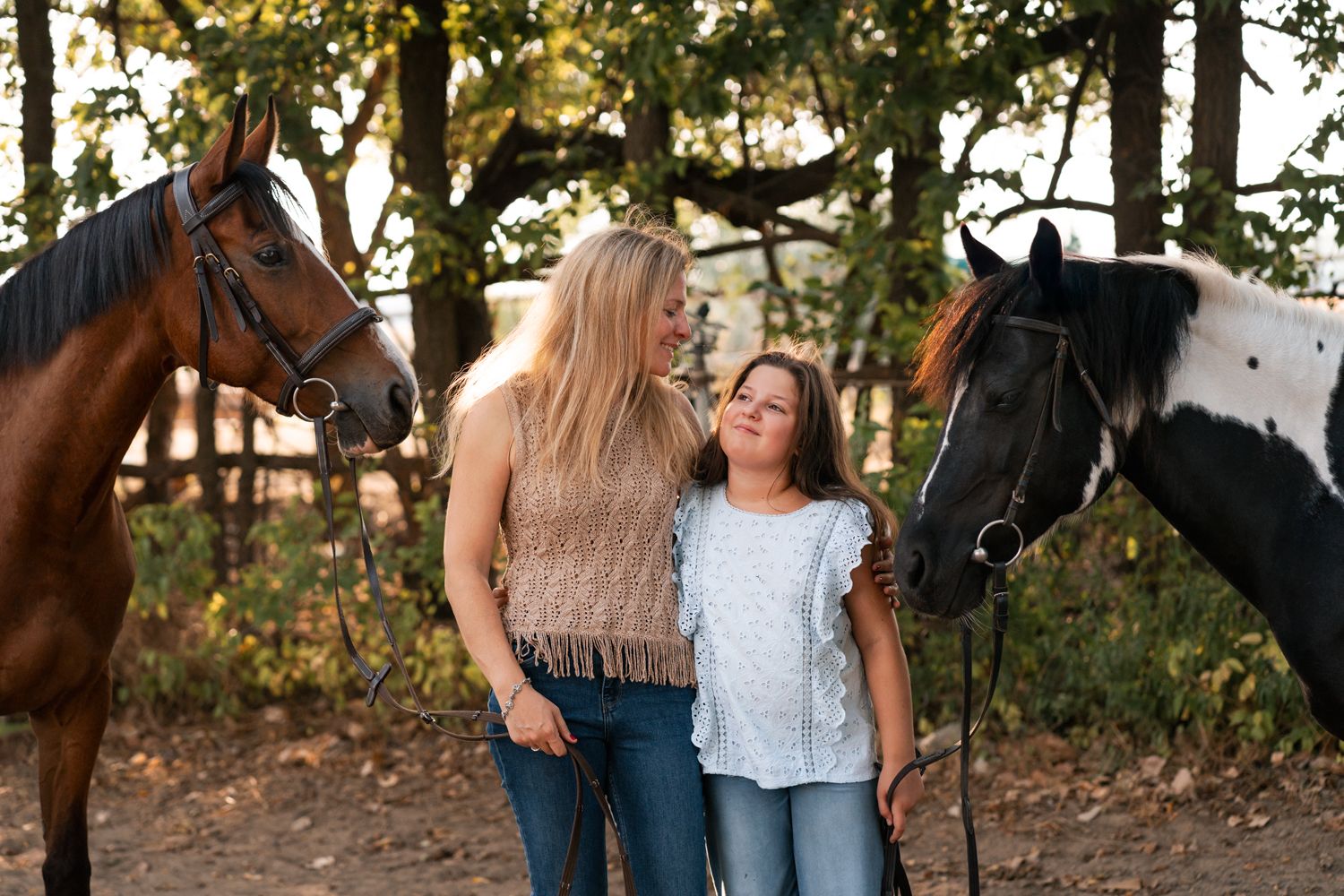 mom and daughter stand between a brown and black horse outdoors in a wooded setting during golden hour family photoshoot in Sydney Cattai