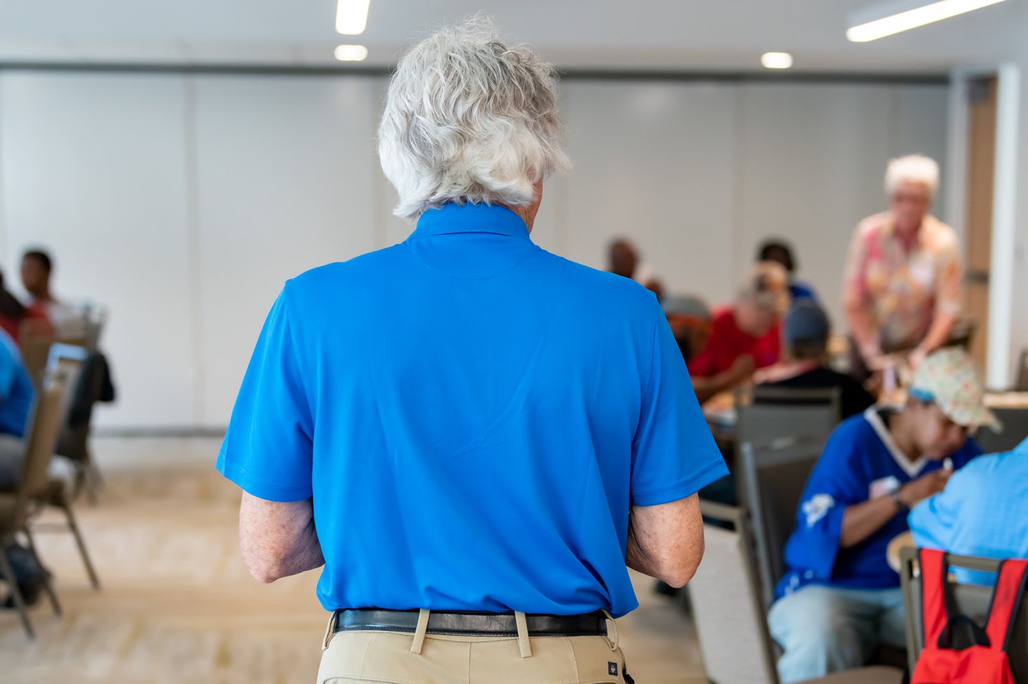 Person in bright blue polo shirt seen from behind speaking to seated audience members in meeting room setting.