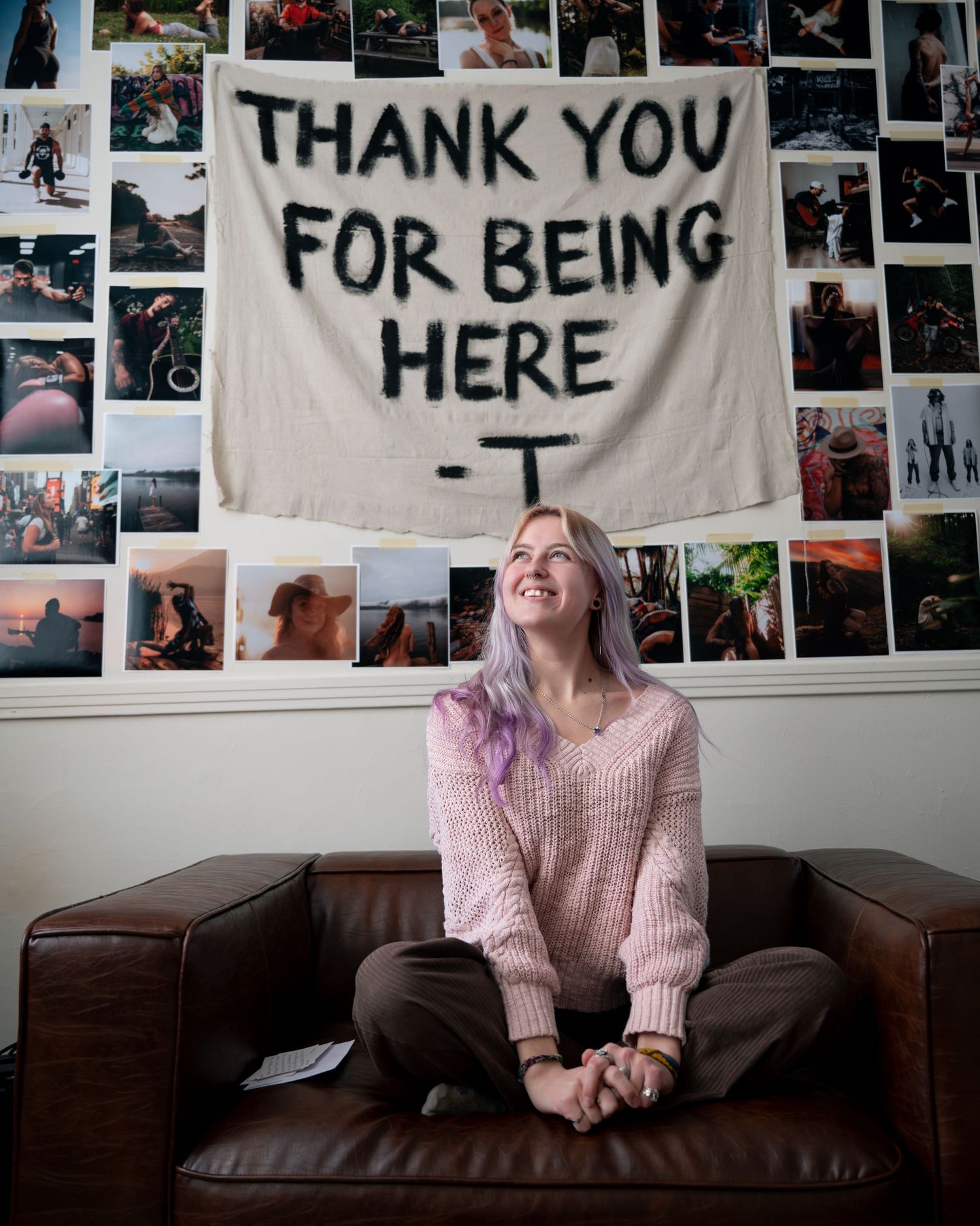 A person in a pink sweater sits on a brown leather couch, with a wall of photos and a 'THANK YOU FOR BEING HERE' banner behind them.