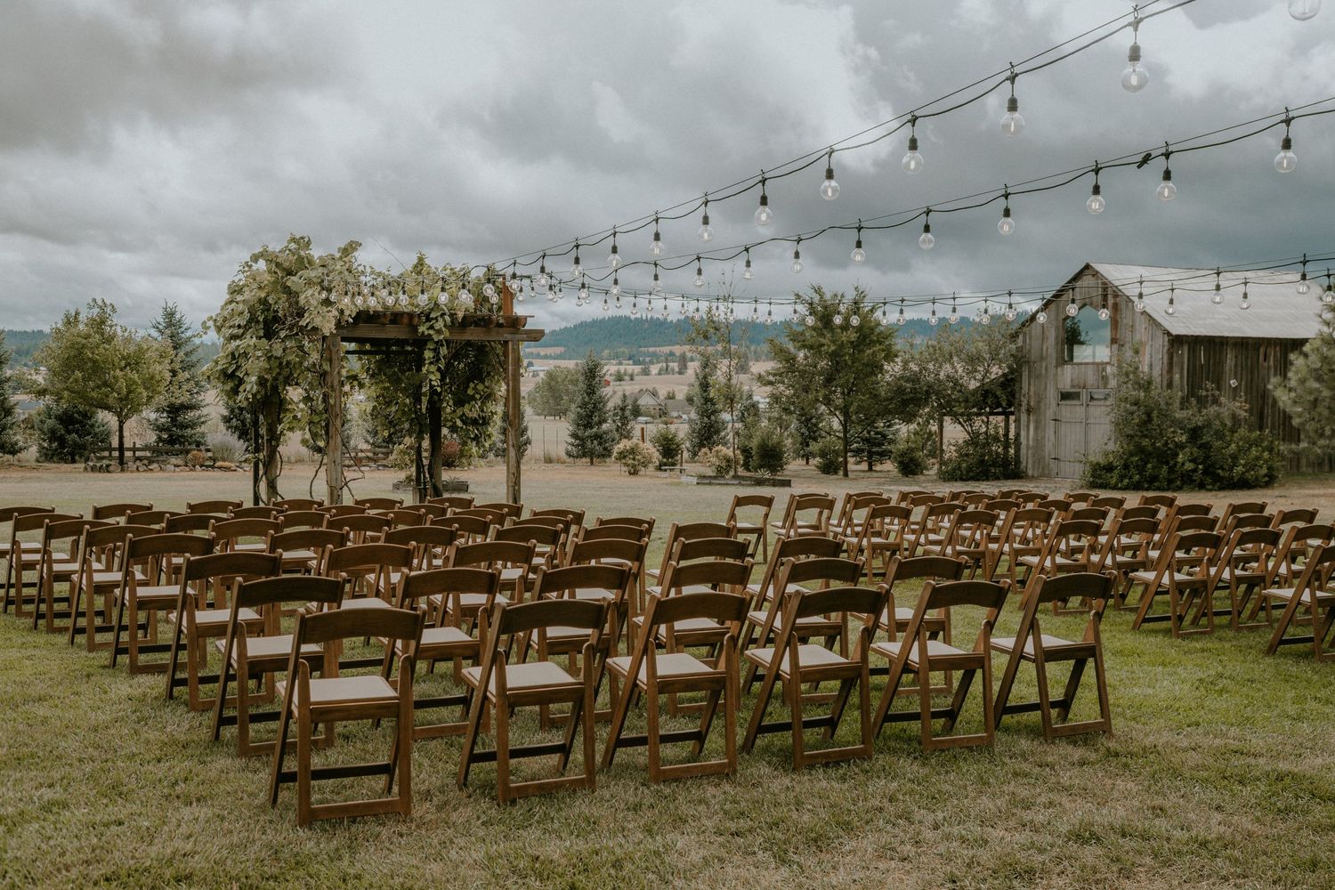 Wooden folding chairs arranged in rows for an outdoor wedding ceremony with string lights and rustic barn in background.