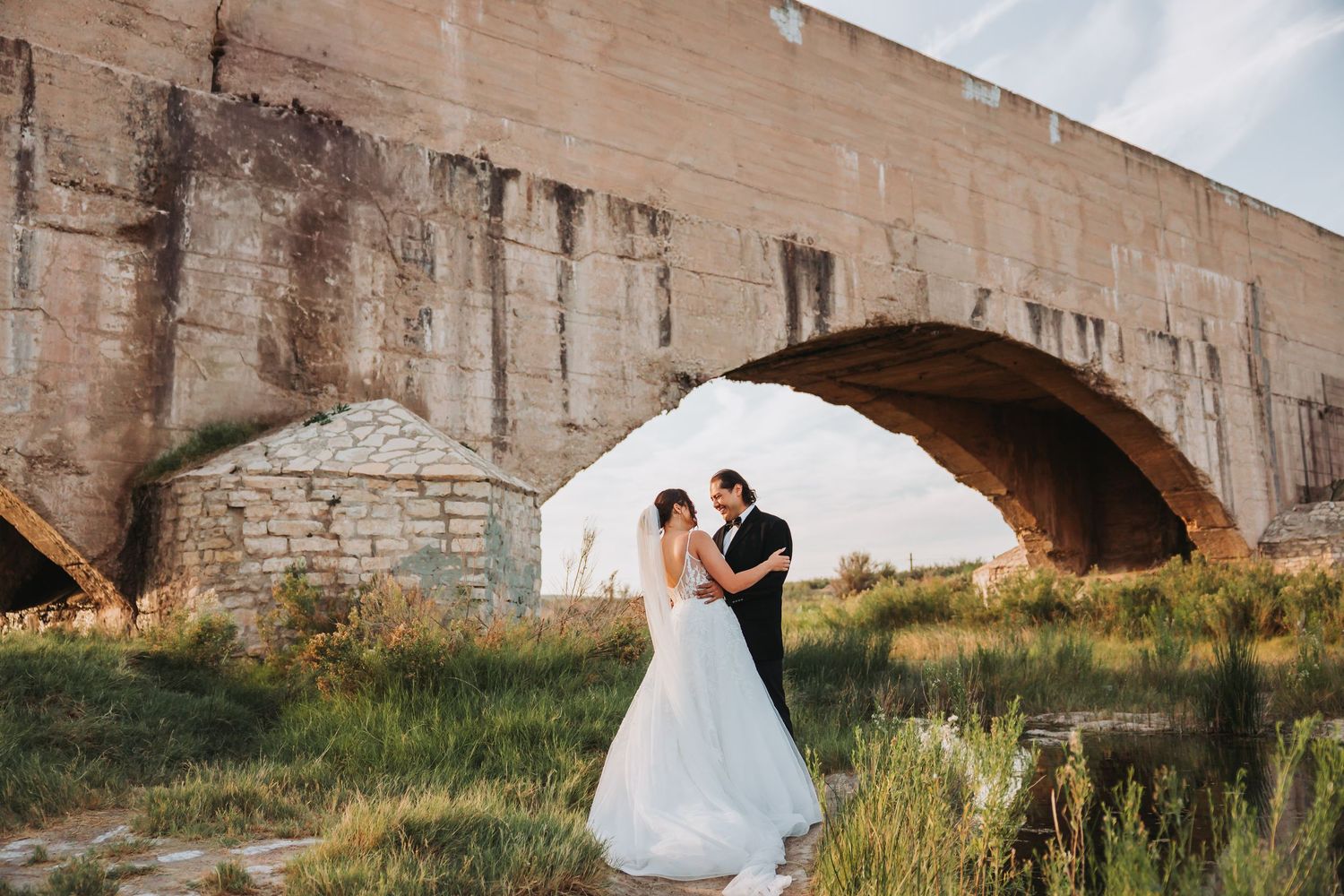 Elegant wedding couple embraces near a historic stone bridge during golden hour sunset.