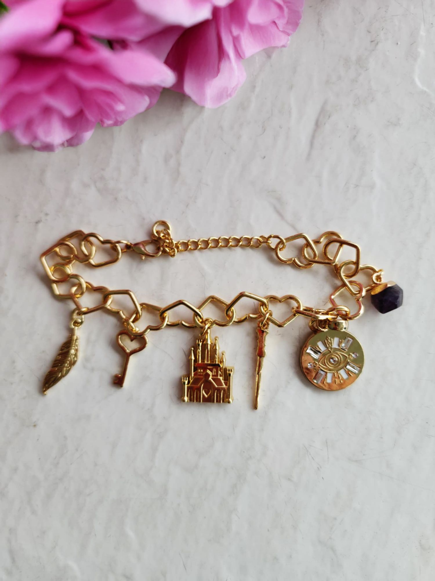 A delicate gold charm bracelet with vintage-inspired pendants including a clock, key, and house against pink flowers.