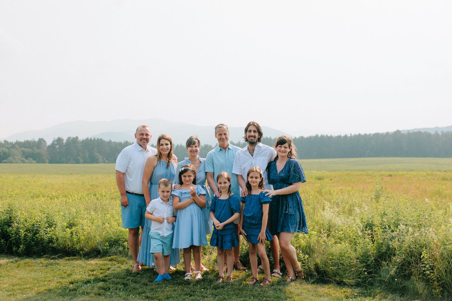 A group of people wearing coordinated blue outfits pose together in a sunny meadow with green fields in the background.