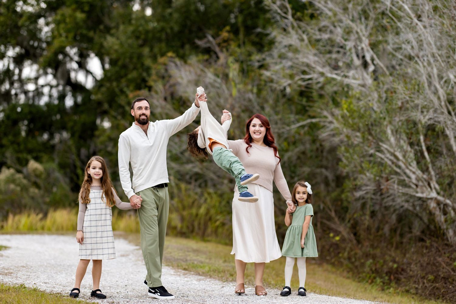 A couple and child hold hands and walk together through a wooded outdoor setting on a sunny day.