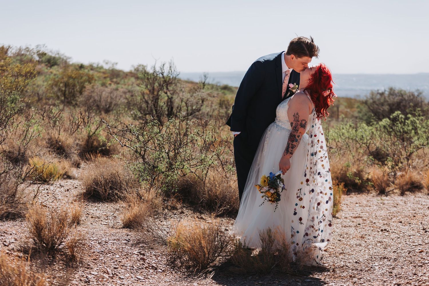 A couple embraces in a romantic moment on a desert path surrounded by desert brush and distant mountains.