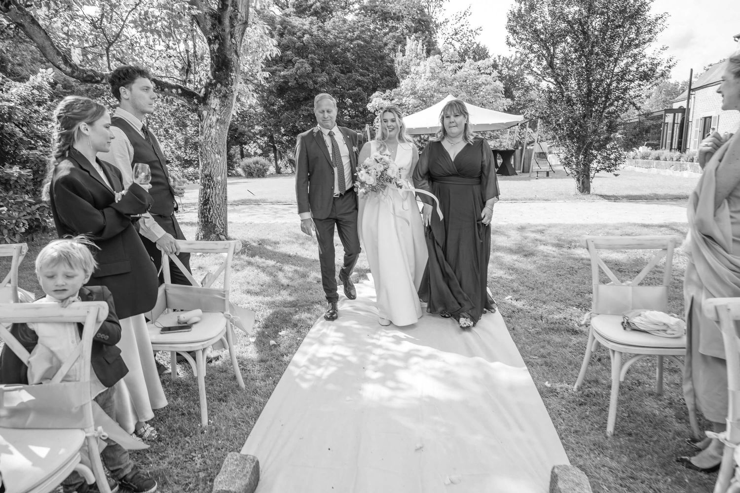 A beautiful outdoor wedding ceremony unfolds on a white-carpeted aisle surrounded by chairs and serene trees.
