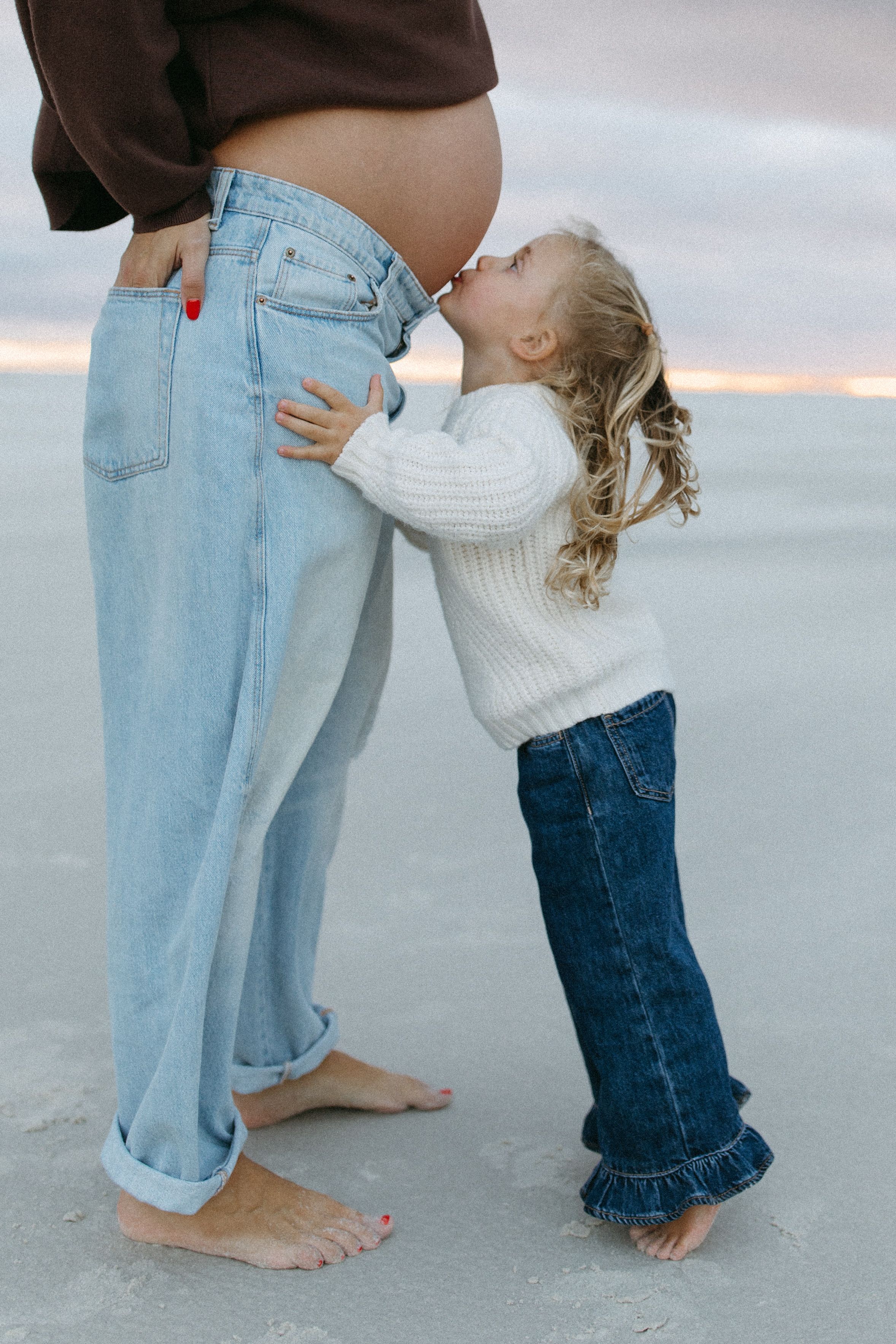 A child in a white sweater hugs an adult wearing jeans and a brown top on a beach.