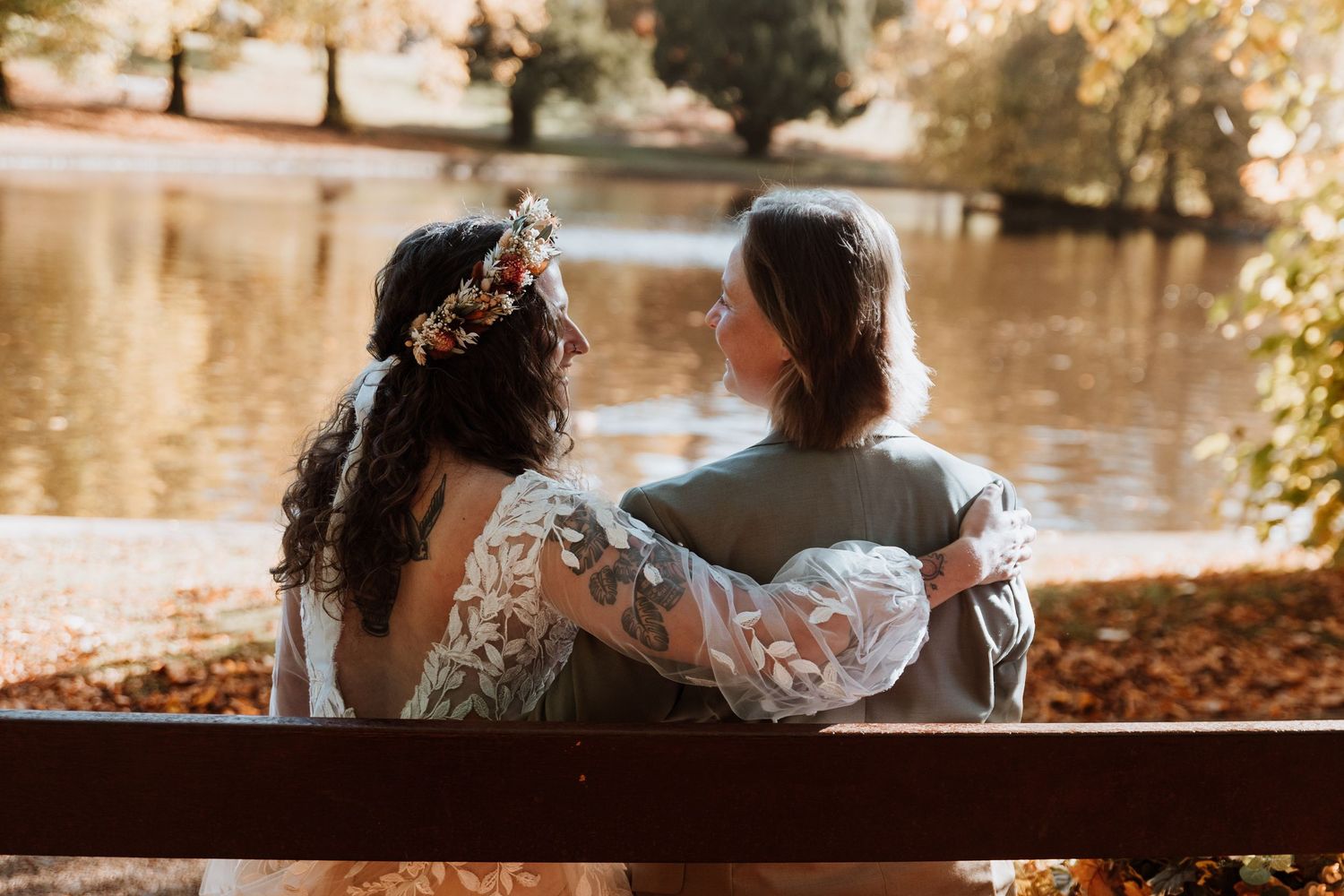 Two people sit close together on a wooden bench by a tranquil pond surrounded by autumn foliage.