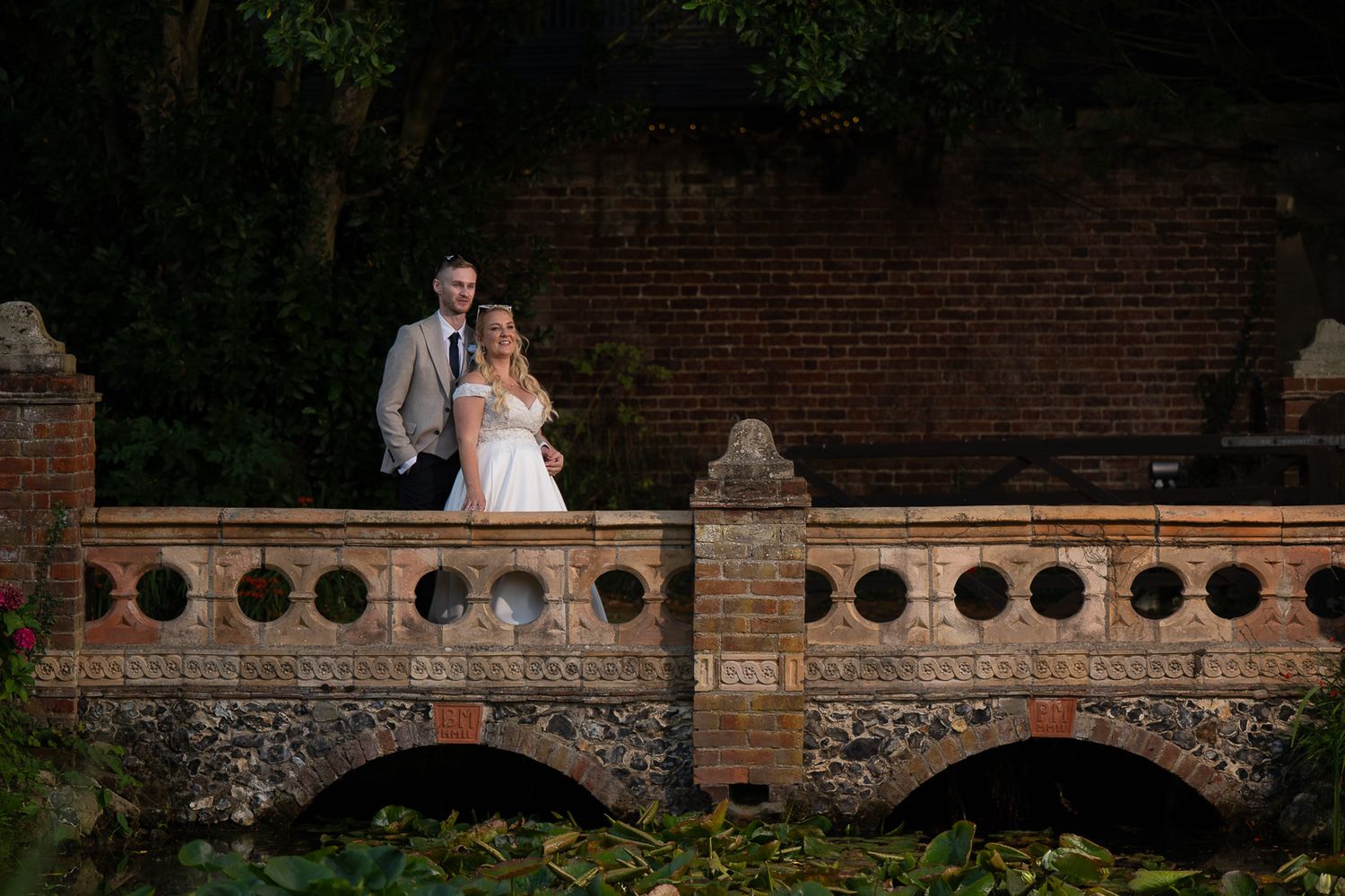 A couple in wedding attire sits together on a rustic brick wall surrounded by greenery at dusk.