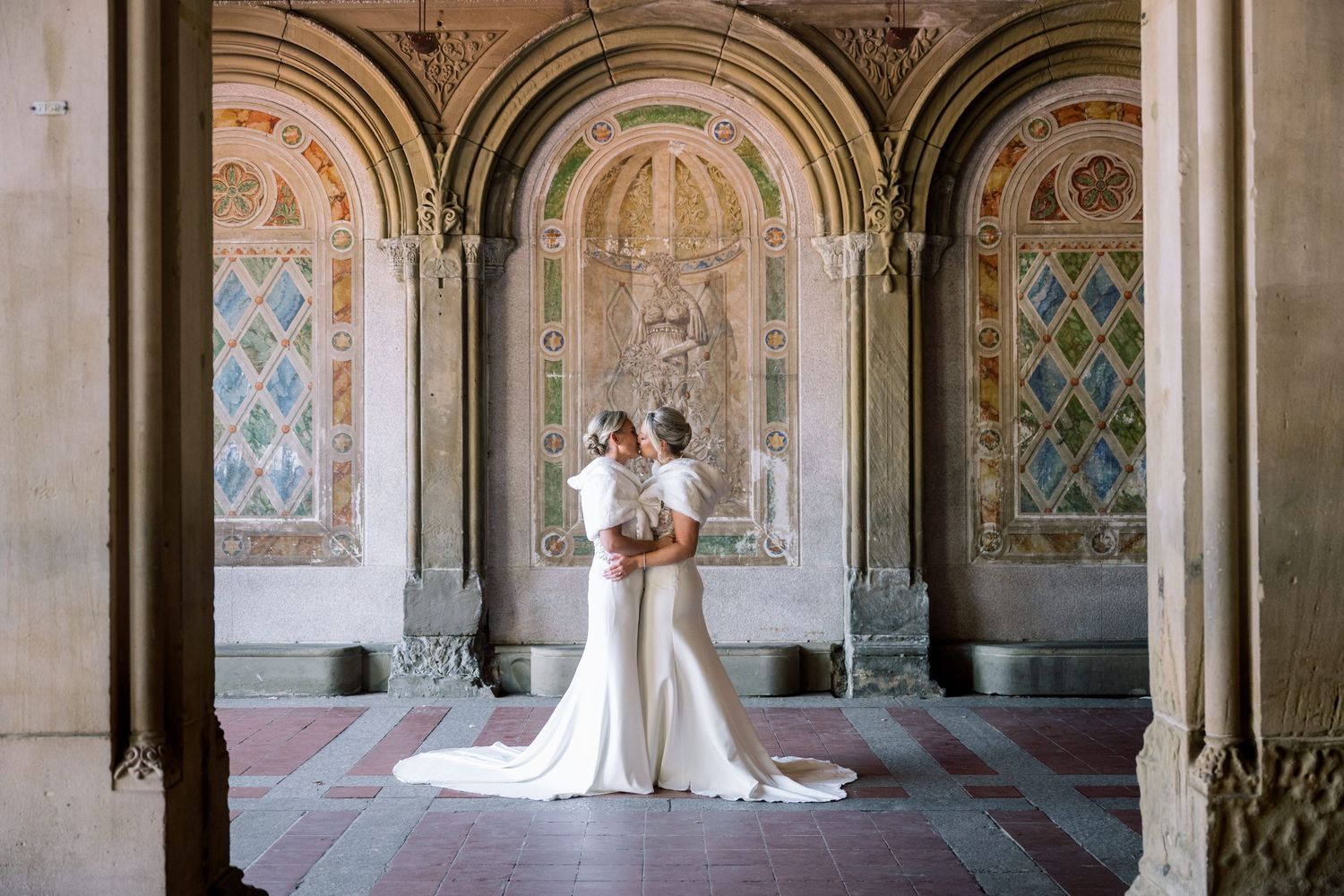 A couple poses by a tall fountain surrounded by manicured hedges and gardens in an elegant outdoor setting.