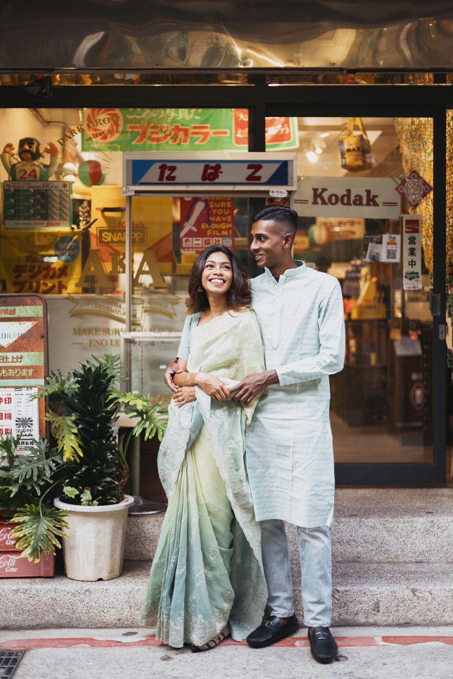 Indian couple in traditional attire smiling outside a retro shop with Kodak signage.