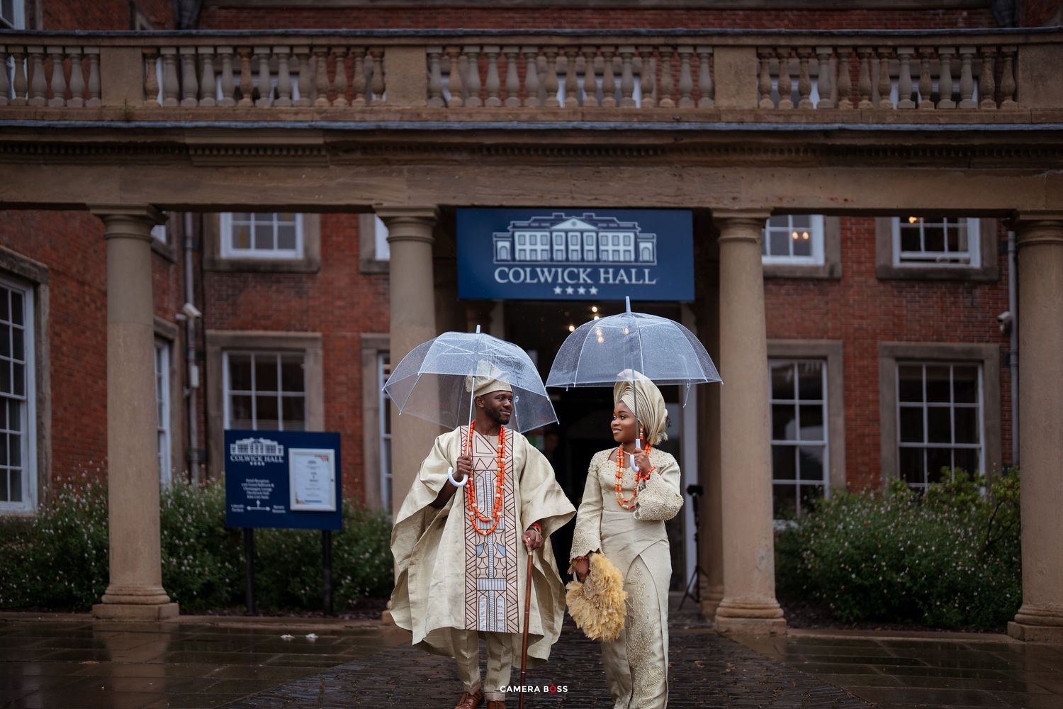 People walk with umbrellas on a rainy day outside a brick building with columns and arched windows.