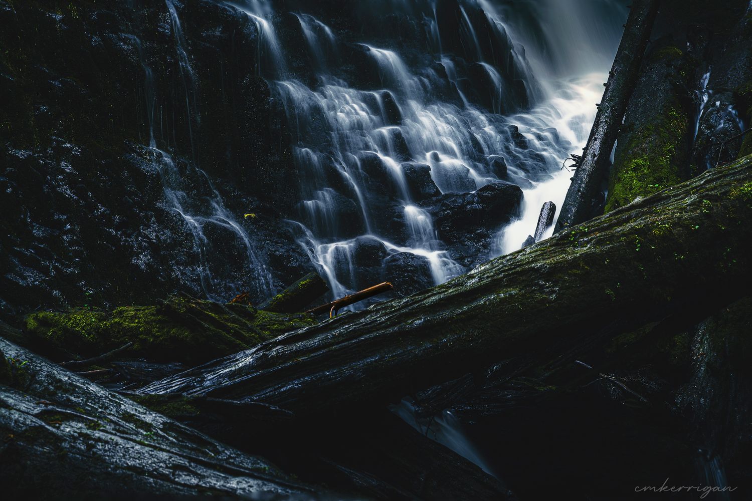 A dramatic waterfall cascades down dark rocky cliffs surrounded by lush forest in moody lighting.