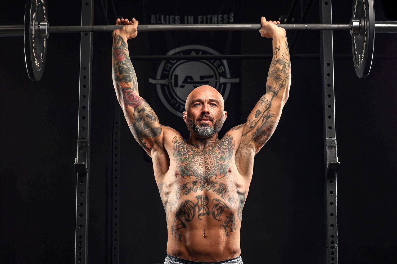 A muscular person with tattoos performs an overhead lift exercise in a gym setting. Professional fitness photoshoot by Boswell Photography.