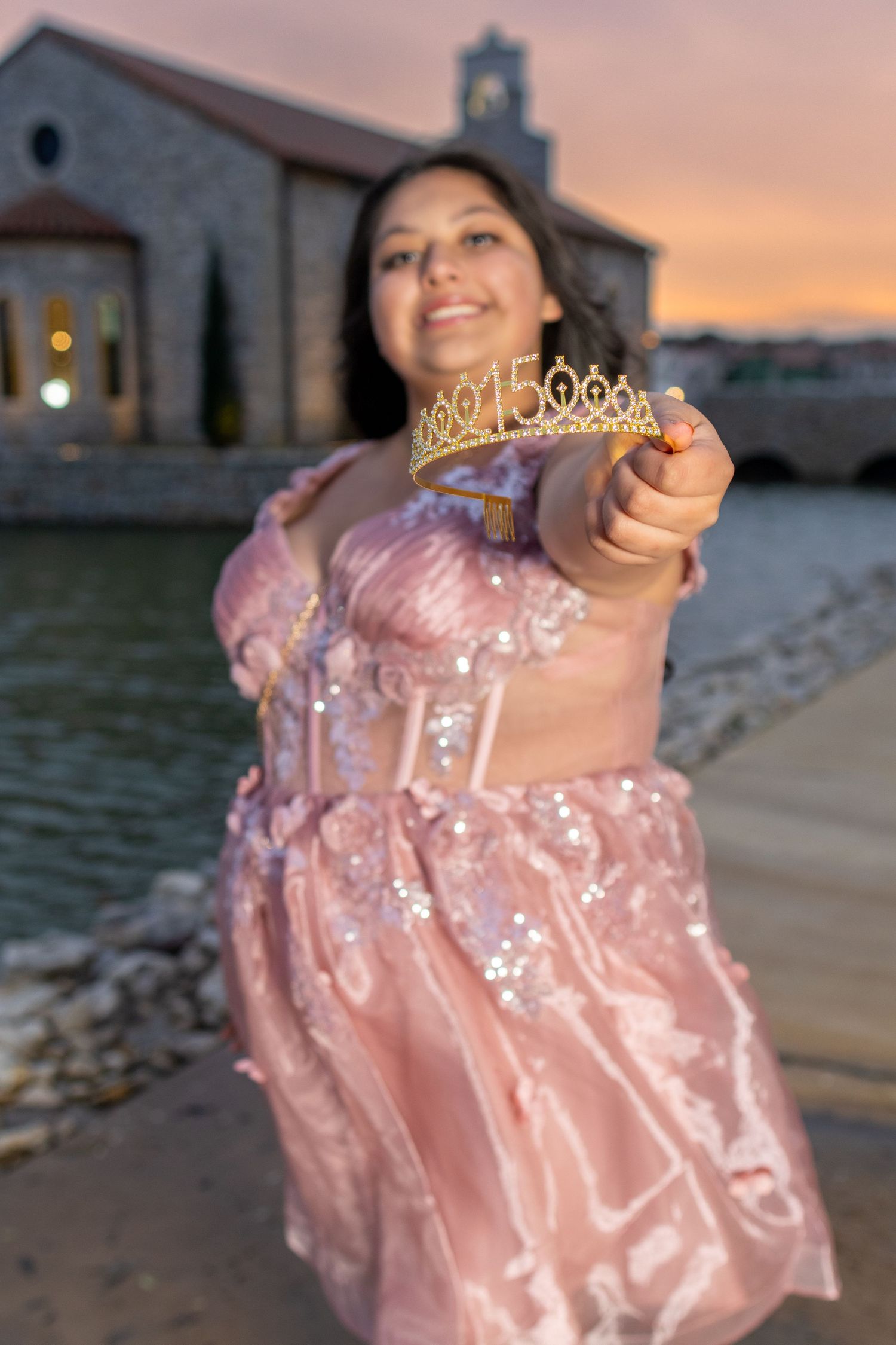 A person in a pink sparkly dress twirls on a beach at sunset with a lighthouse in the background.