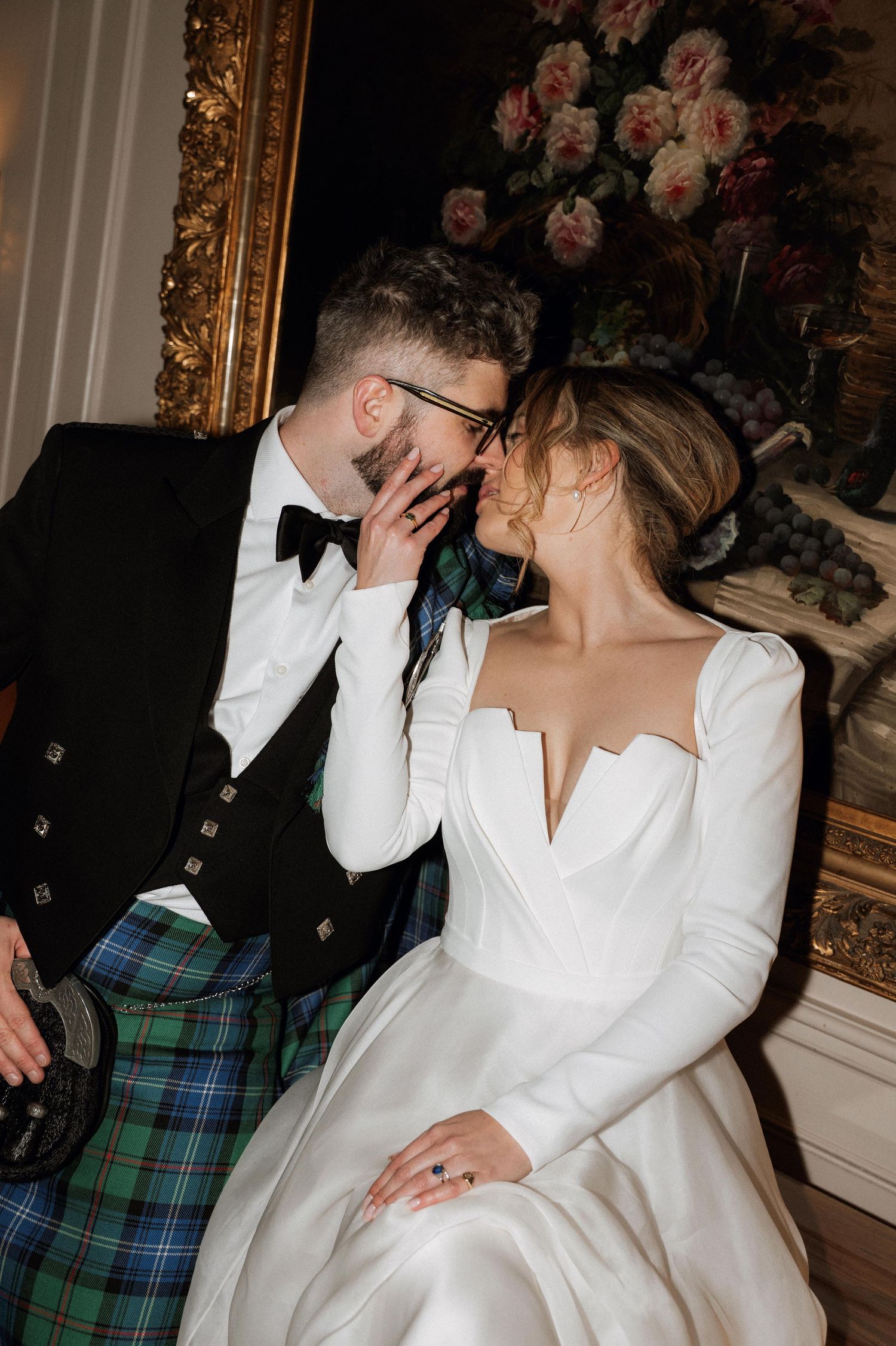 A couple in formal attire share an intimate moment during a wedding celebration with floral decor in the background.