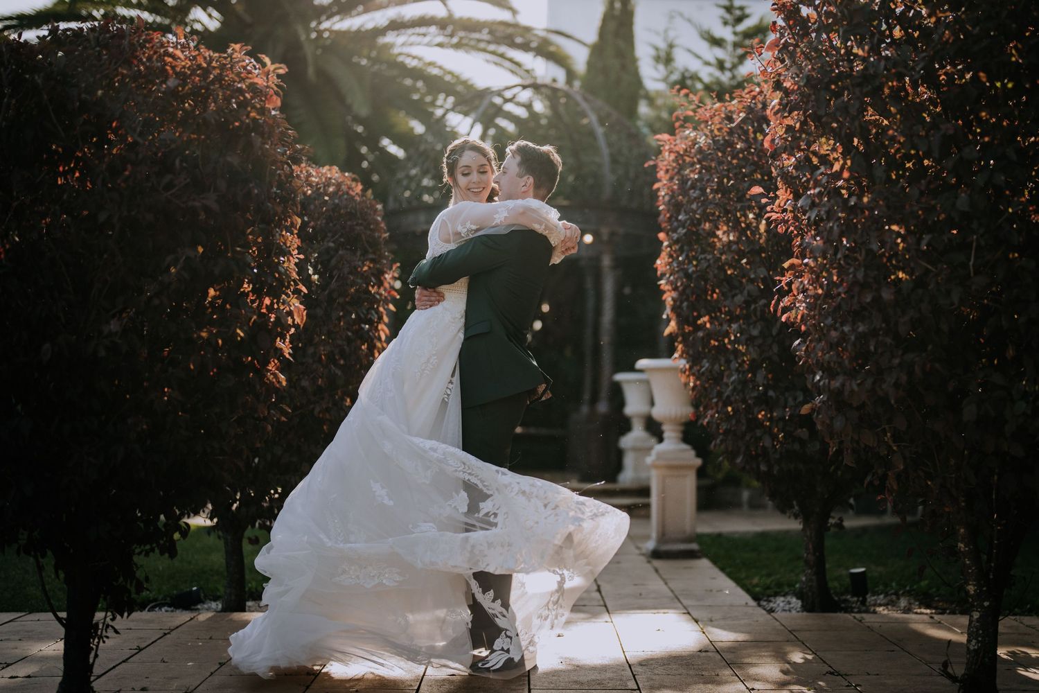Newlyweds share romantic dance moment in garden path surrounded by blooming trees.