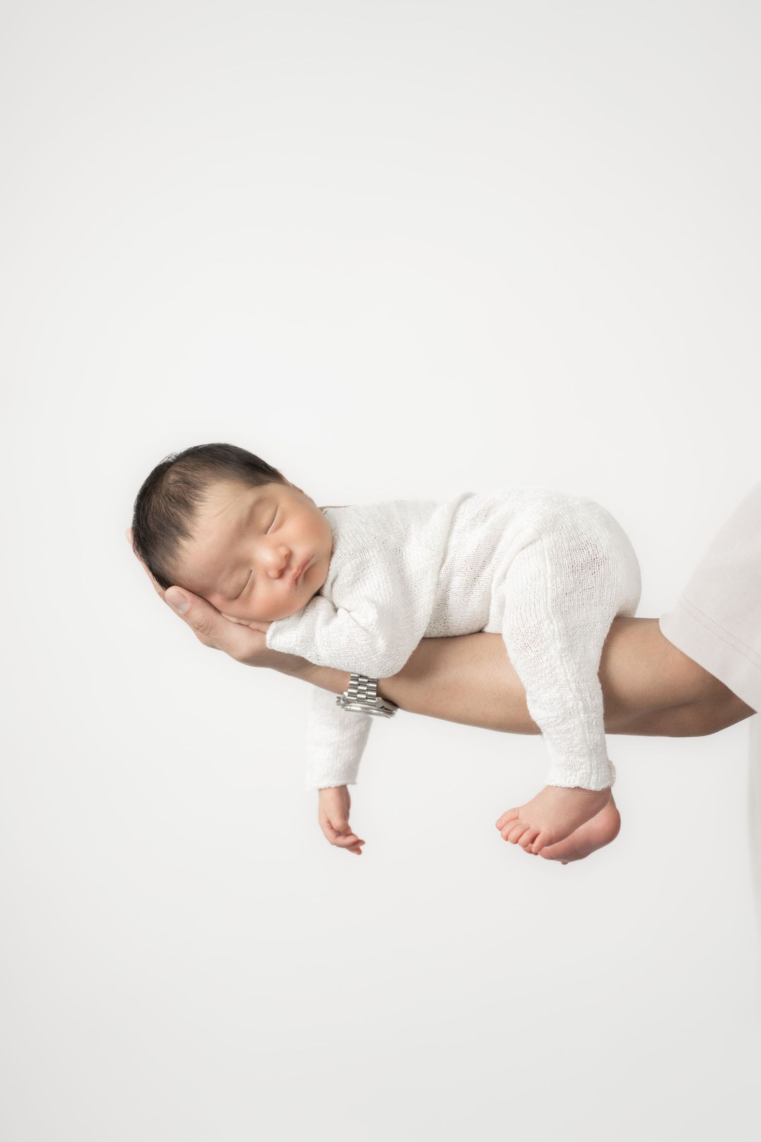 A sleeping baby wearing white clothing lies peacefully on a white background in a minimalist portrait.