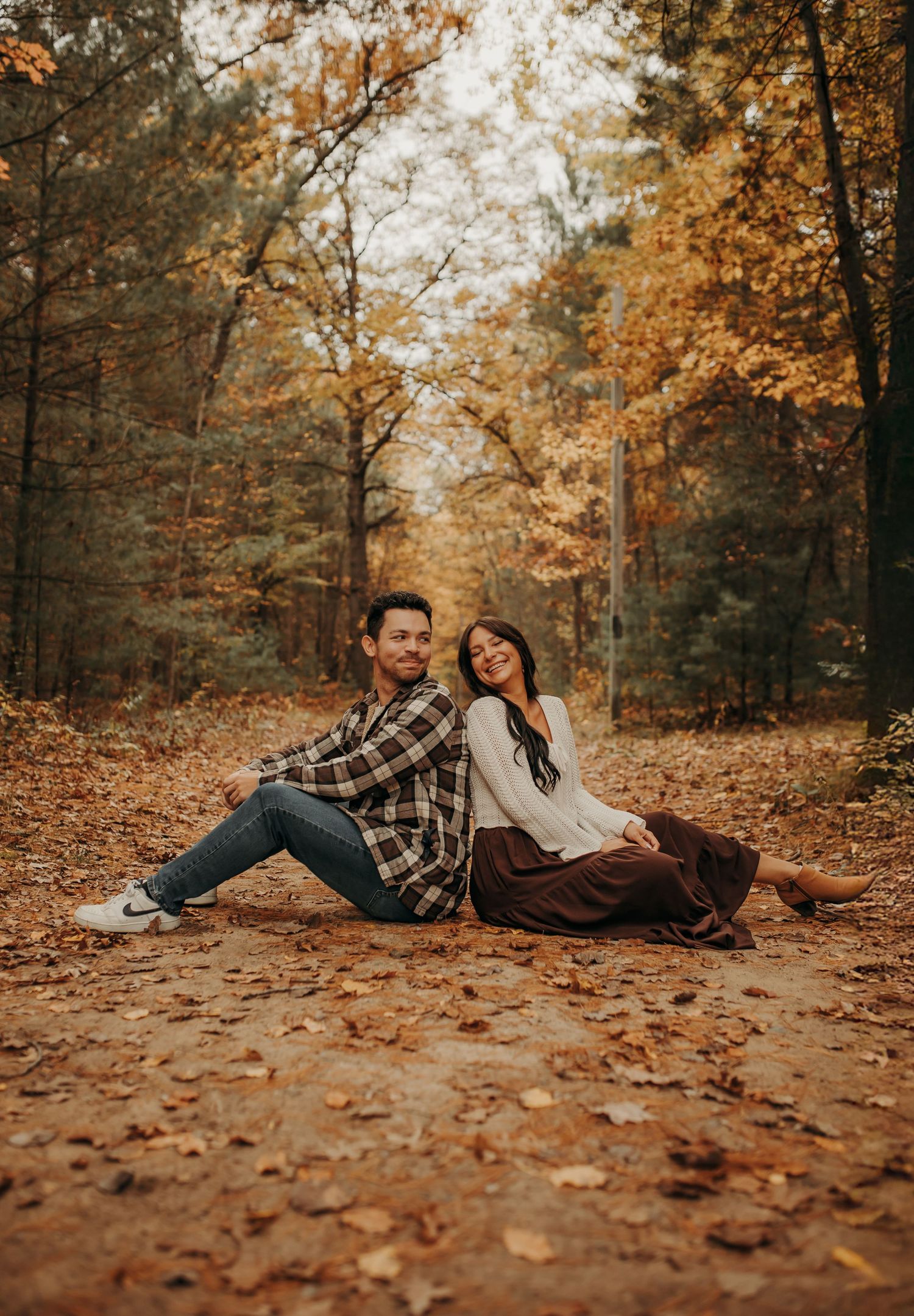 A couple sits back-to-back on fallen autumn leaves in a forest with golden foliage during fall season. Creative Art By Bree LLC