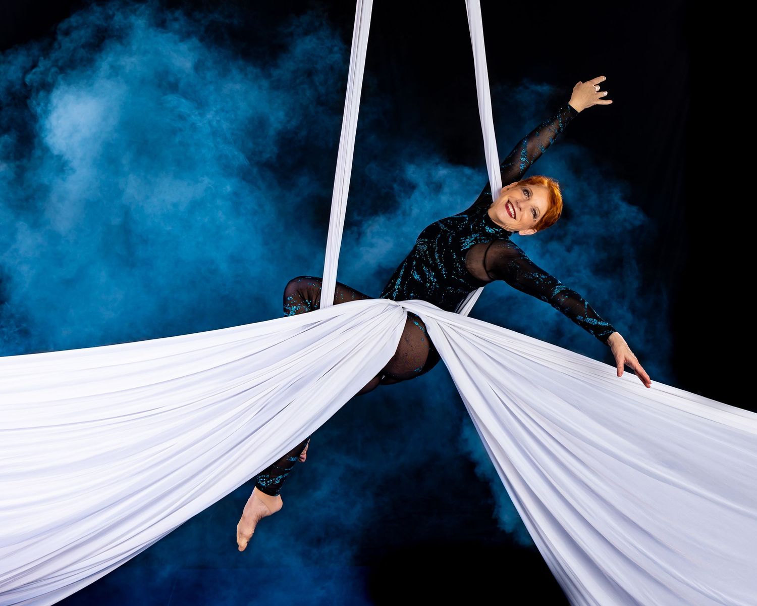 An aerial performer gracefully moves with flowing white fabric against a dramatic blue background in a dynamic dance sequence.