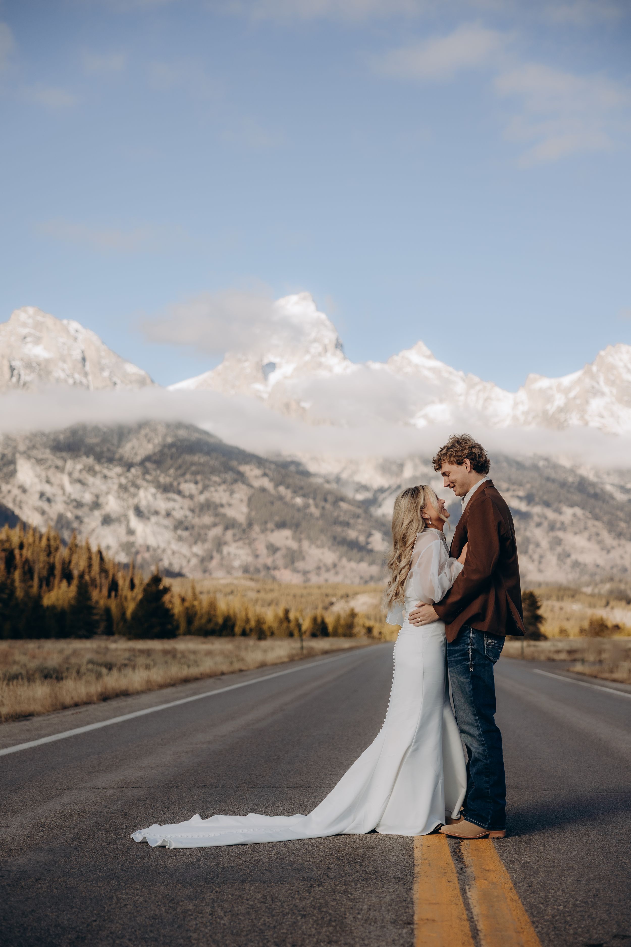 A couple embraces in a golden field with snow-capped mountains and evergreen forests in the background.