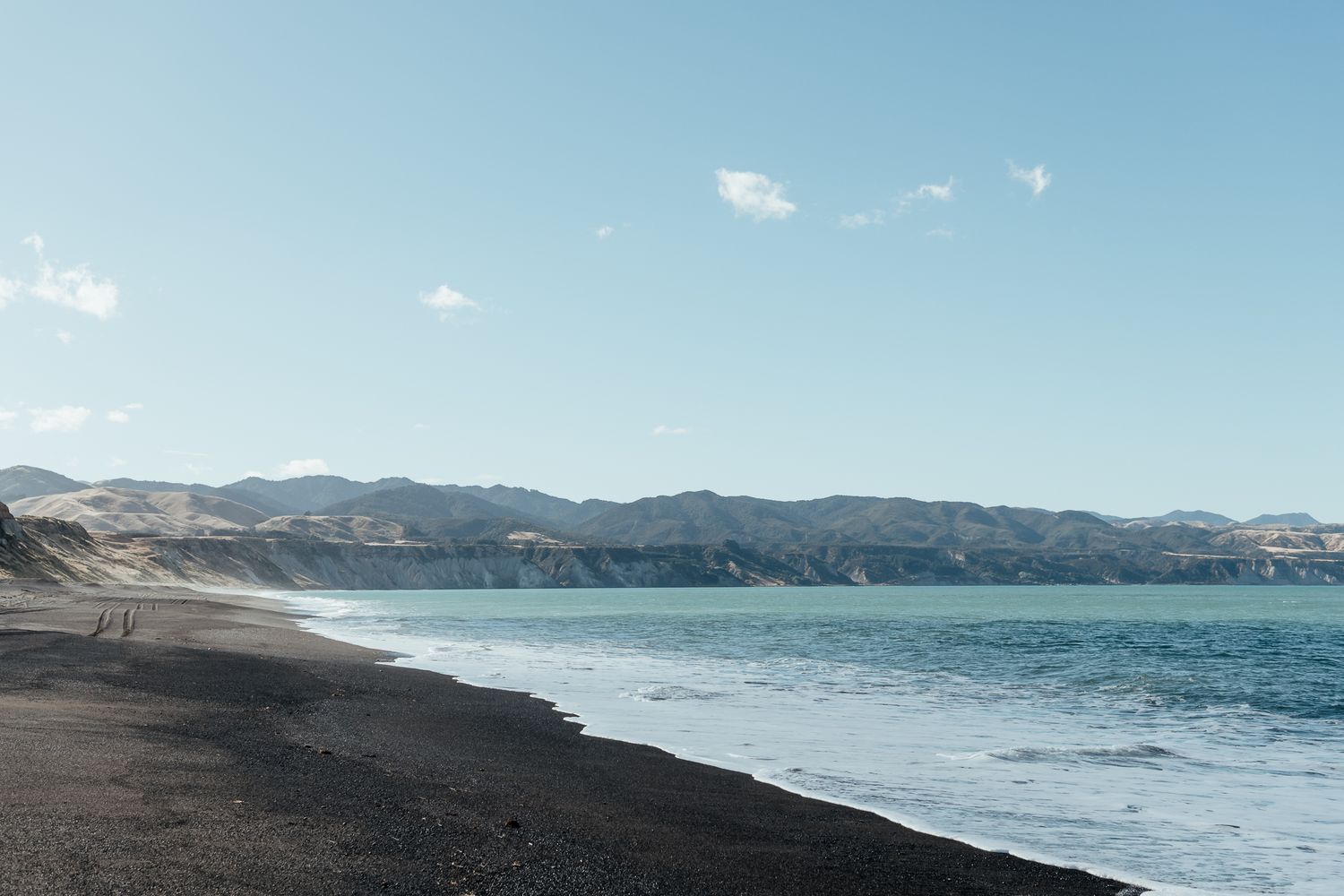 Panoramic view of a Whangaimoana beach Wairarapa with rolling waves and mountains in the distance under a blue sky by August and Vine