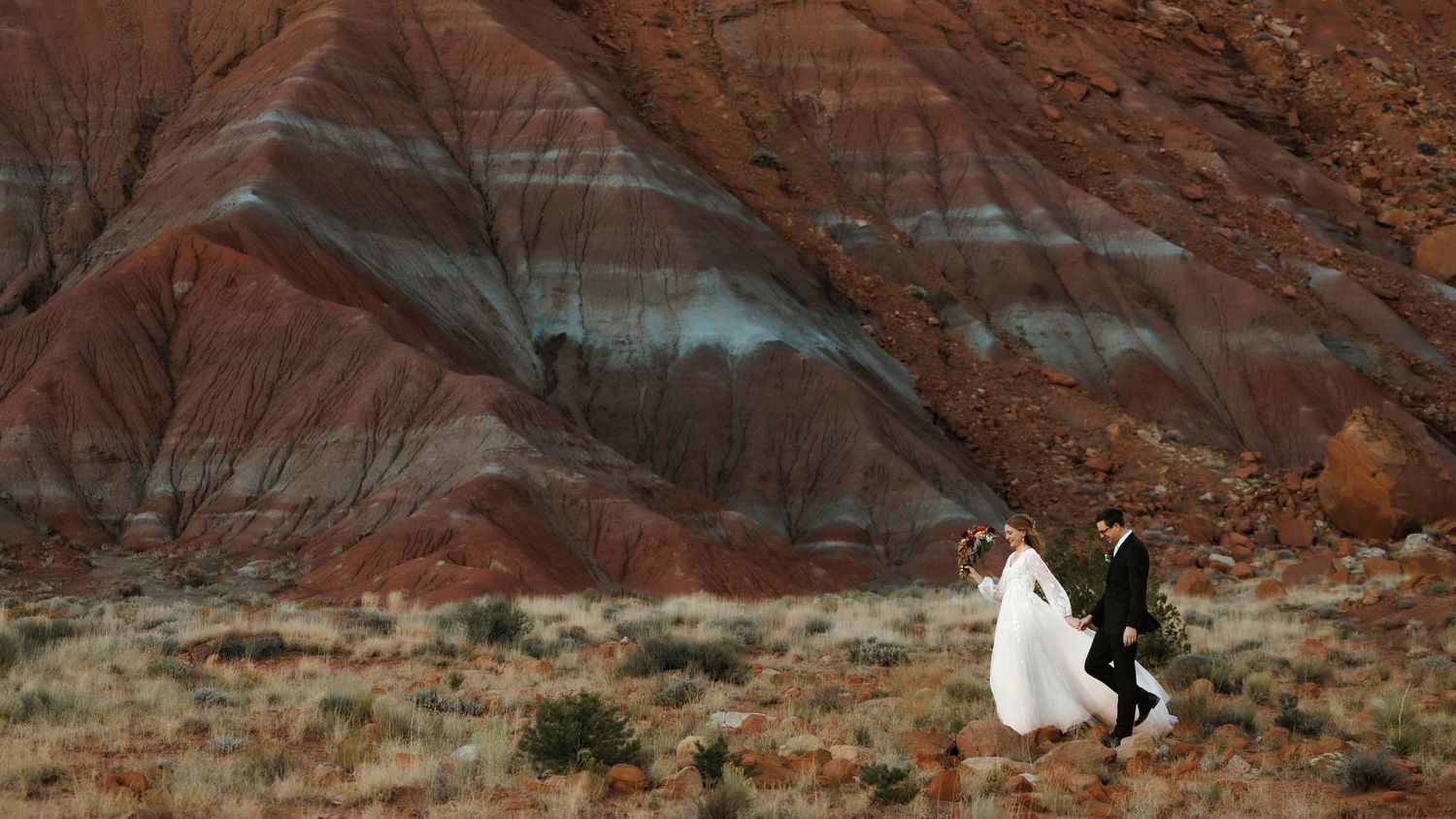 Bride and groom walking hand in hand against dramatic red rock desert landscape.