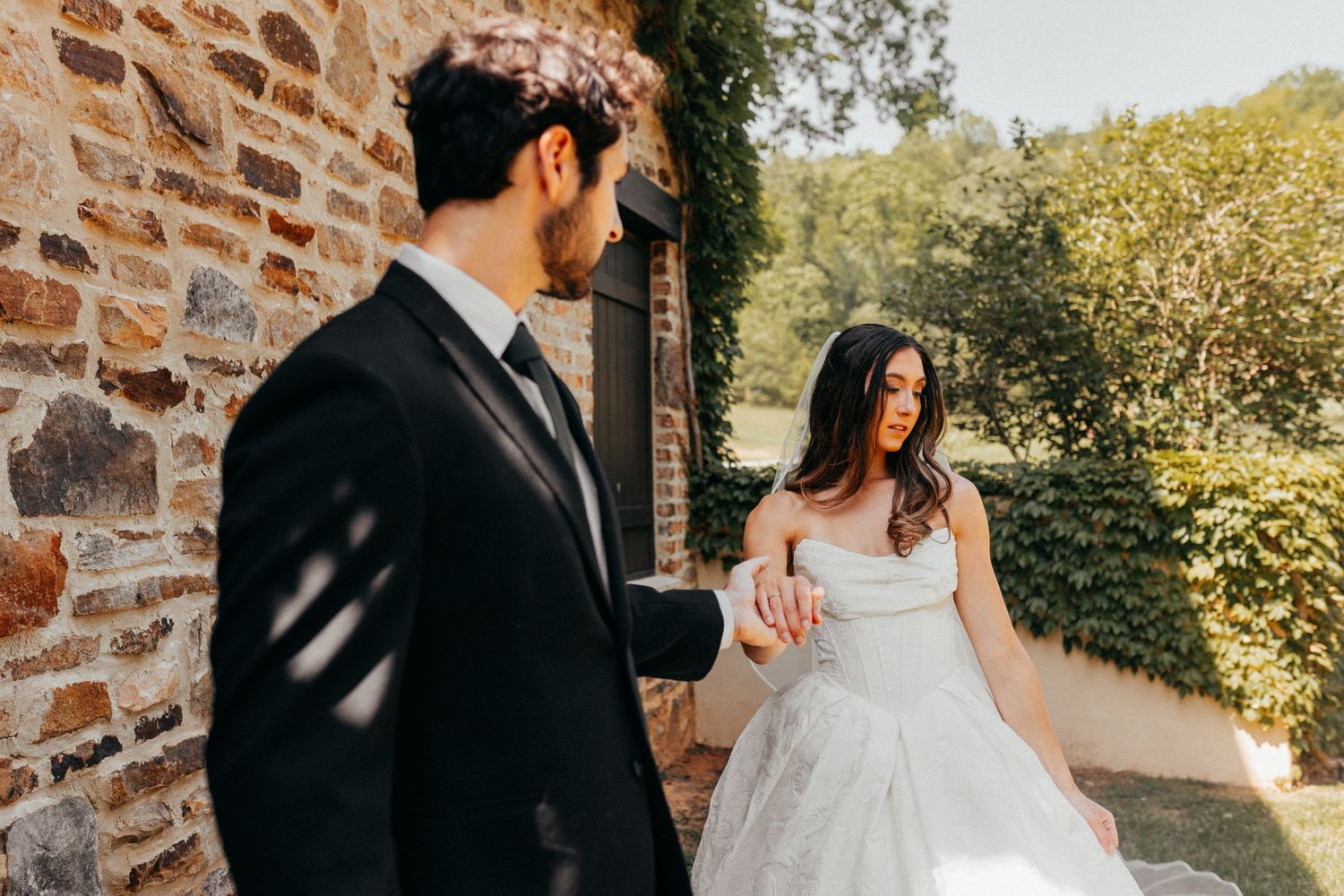 A couple in wedding attire hold hands while walking along a rustic stone wall on their sunny wedding day.