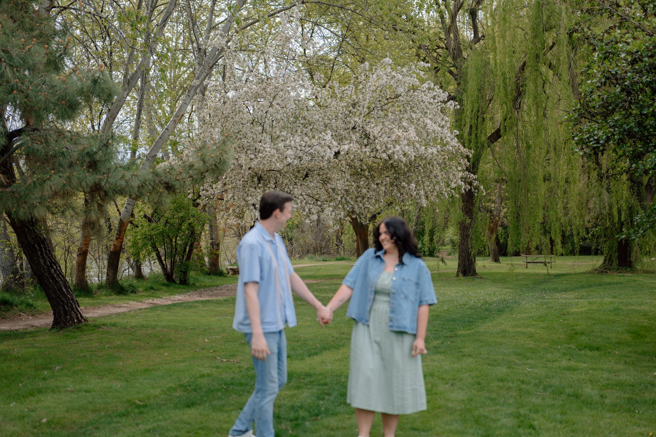 A couple holds hands while walking through a spring garden with blooming cherry trees and willow branches.