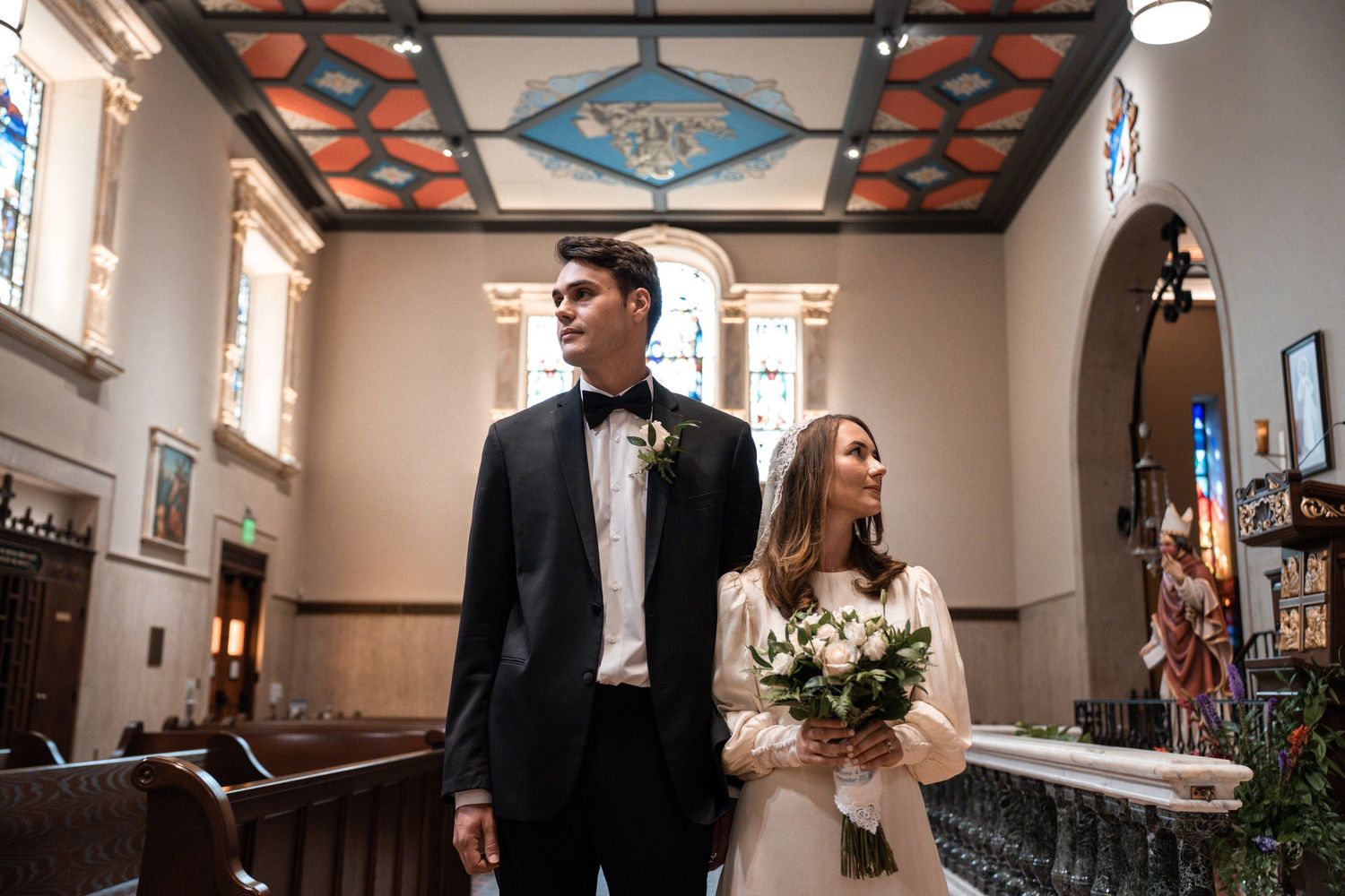 A wedding ceremony takes place in a church with white walls and stained glass windows as the couple stands at the altar.
