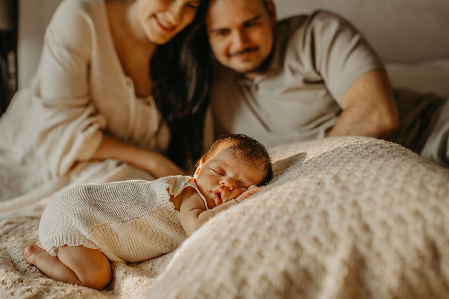 A sleeping newborn baby in white clothing lies on a textured cream blanket while parents watch from above.
