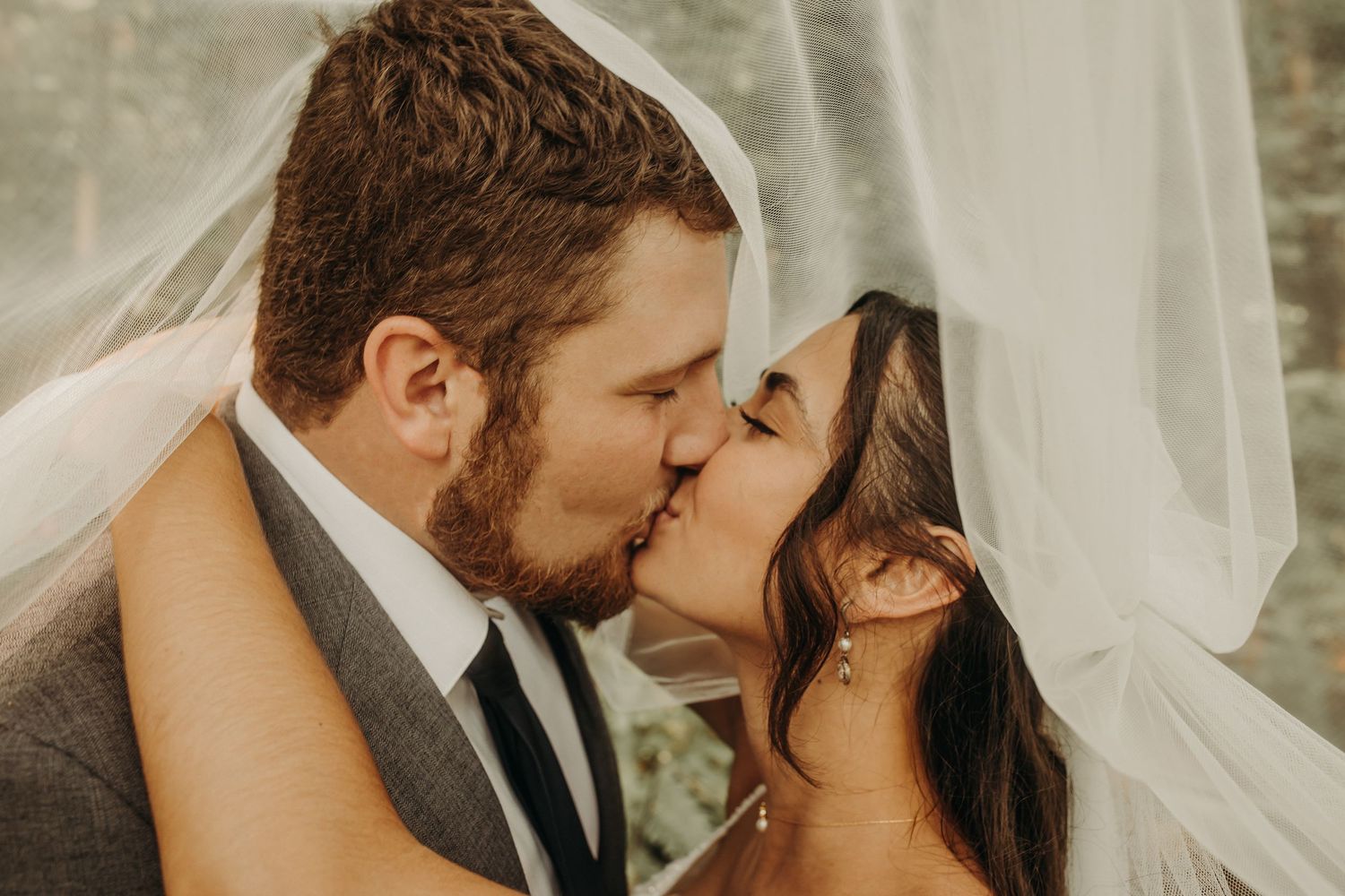 Romantic wedding couple shares tender kisses under flowing white veil in outdoor setting.