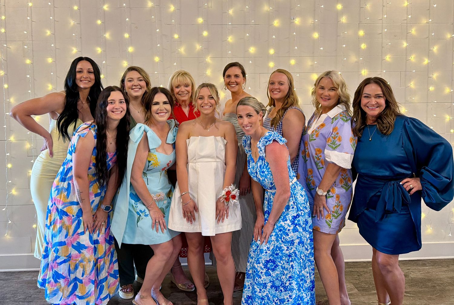 Group of women in colorful dresses posing together at a festive event with twinkling lights backdrop.