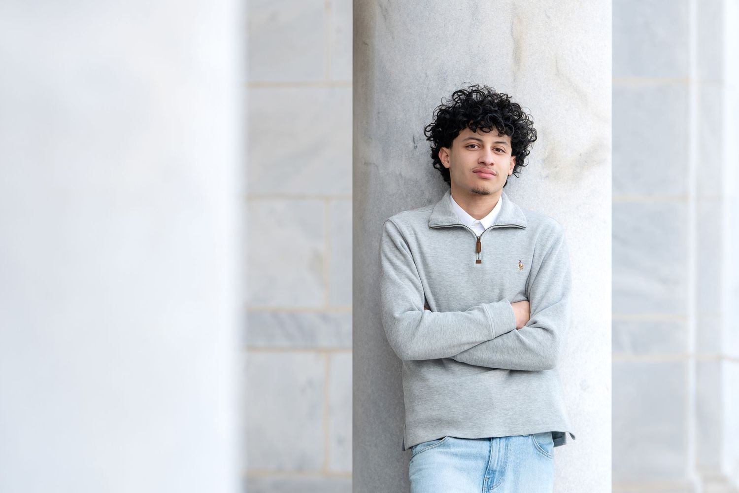 Teen boy with curly hair in gray pullover leaning against marble columns with arms crossed.
