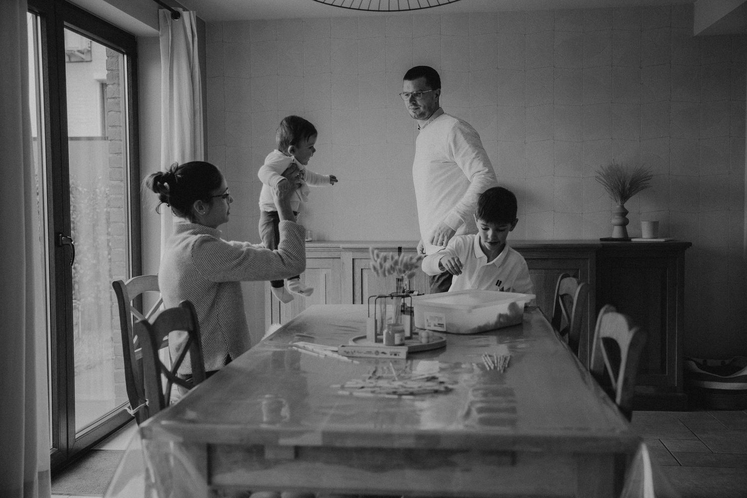 Black and white photo of a family gathered around a dining table in a bright home interior.