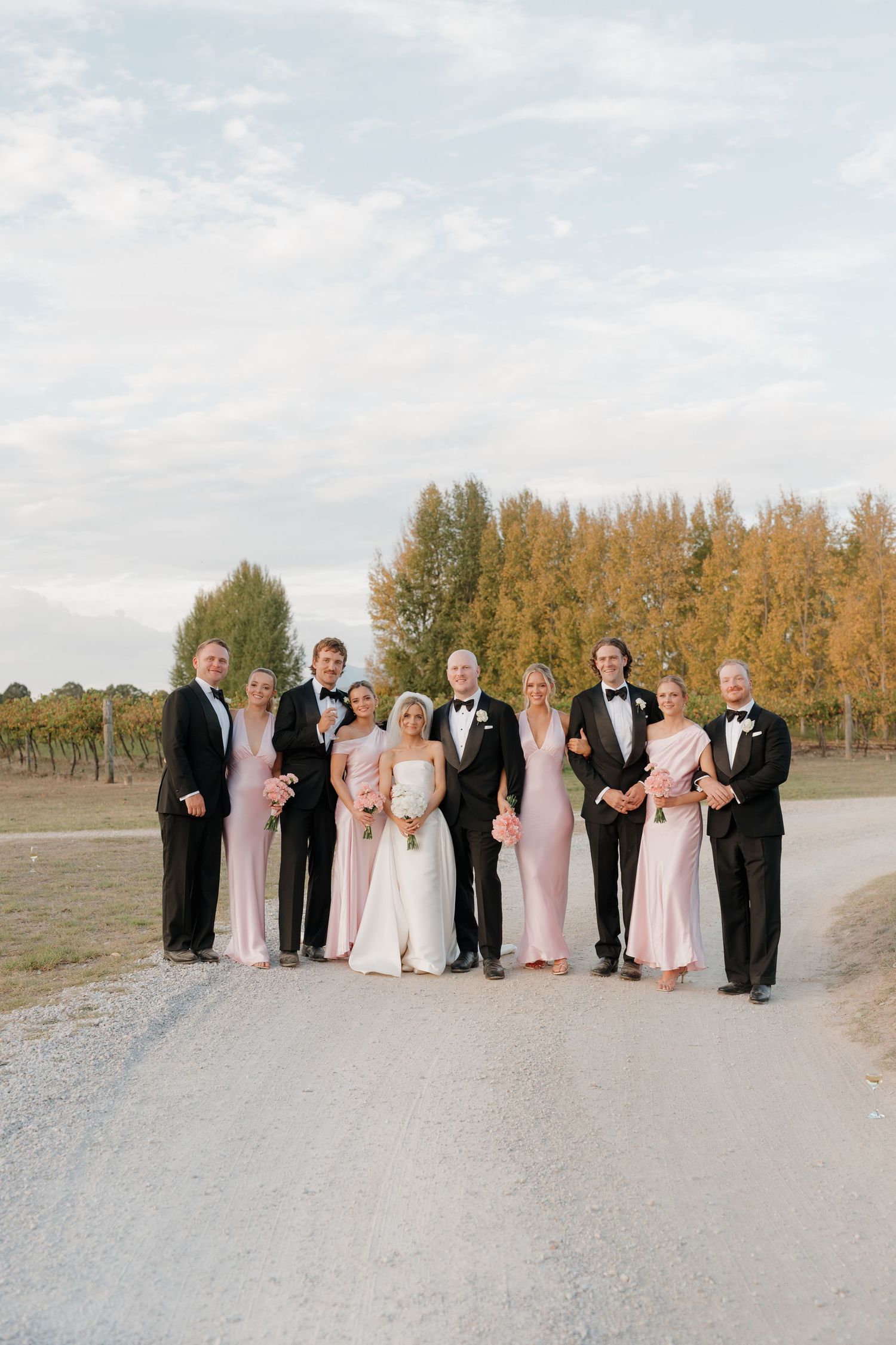 Wedding party in black tuxedos and blush pink gowns posing on a gravel path at a vineyard during golden hour.