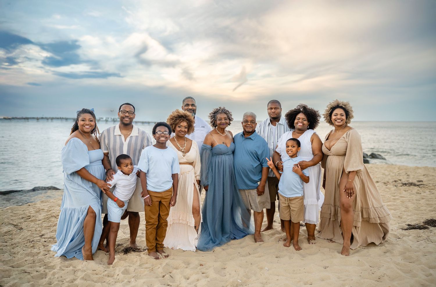 Large Black family posing together on sandy beach at sunset, wearing coordinated white and blue outfits.