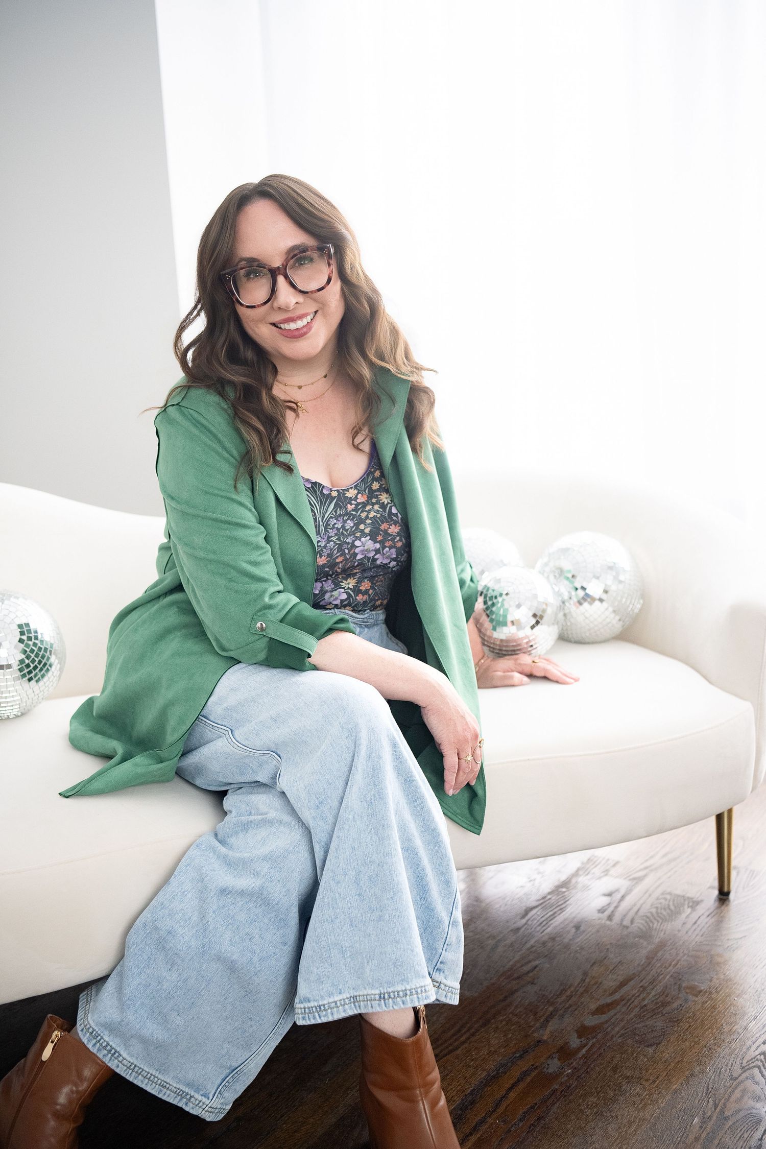 Woman in green blazer, floral top, and wide-leg jeans sitting on white sofa, smiling, wearing glasses.
