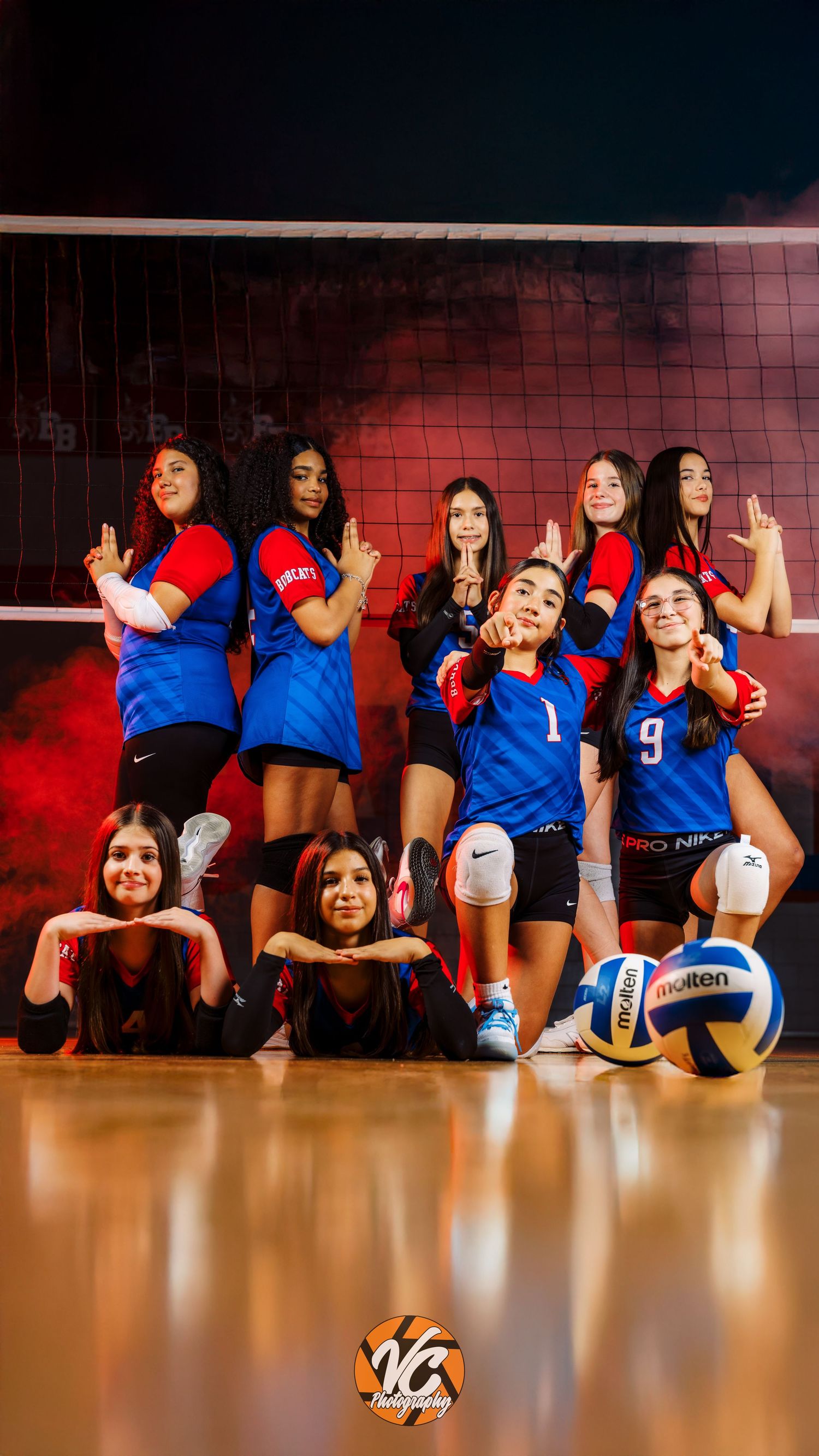 Girls volleyball team posing in red and blue Nike uniforms on a court with a Molten volleyball and dramatic red smoke background.